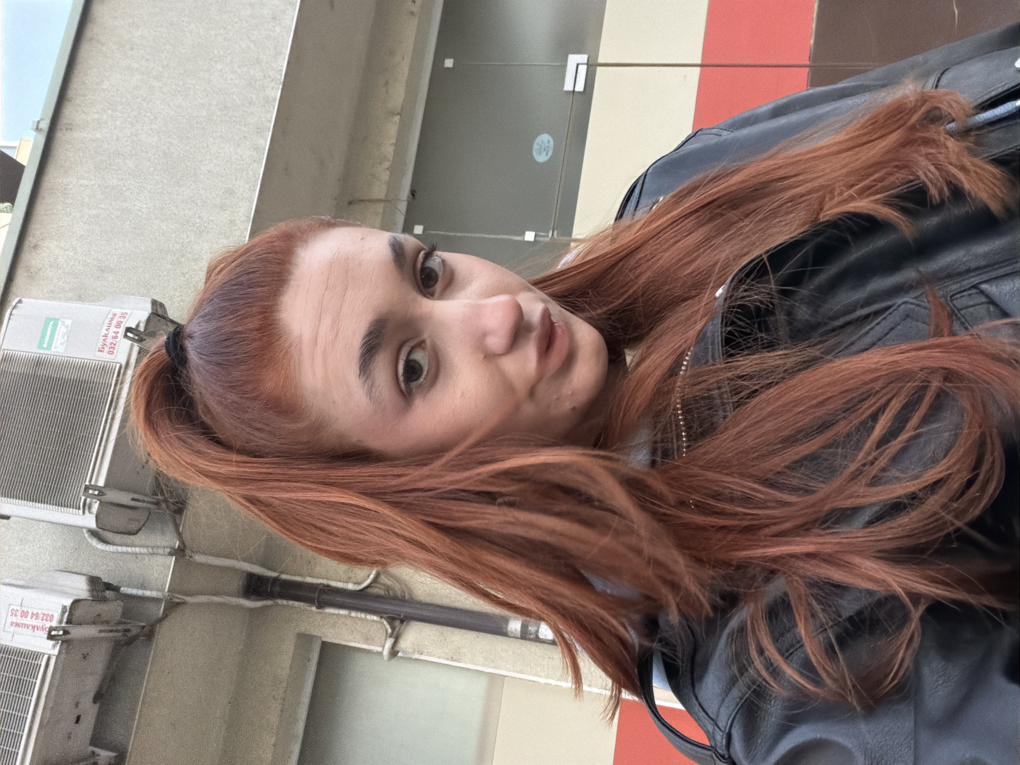 A woman with auburn hair pulled back in a partial ponytail and a black leather jacket takes a selfie outdoors. The background is an urban environment. Visible are the facade of a building with air conditioning units, a glass door, and a red and white stripe. The air conditioning units have writing in a foreign language (likely Bulgarian) and the phone number 032 64 00 35. Given the phone number area code, the location is likely Plovdiv, Bulgaria.