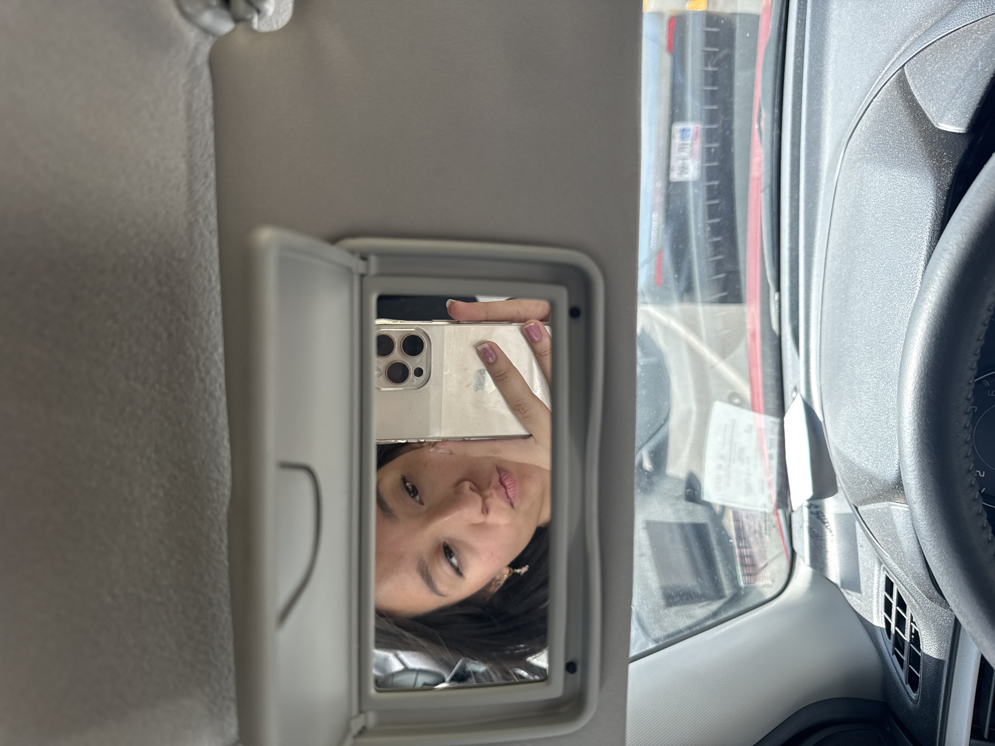 The image shows a person taking a selfie in the mirror of the sun visor of a car. The person is holding a phone with multiple camera lenses. The car is on a road, with a building or structure visible through the windshield. The location in the city of Puebla, Mexico cannot be determined from the image.