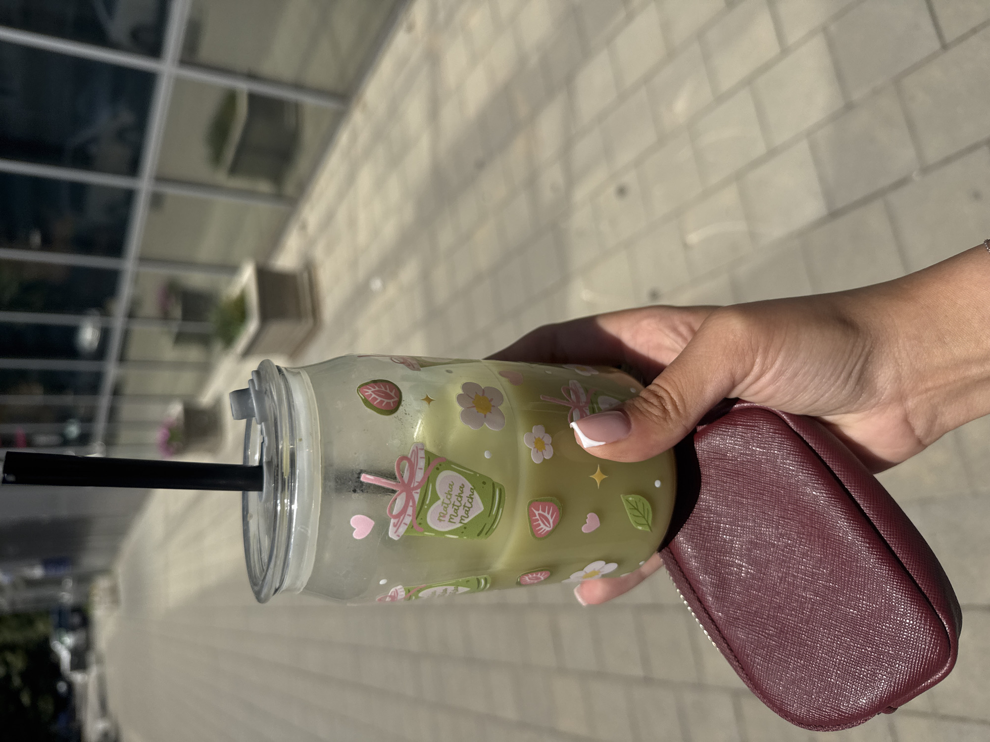 A close-up, eye-level, outdoor shot shows a person's hand holding a clear glass cup filled with a light green beverage. The cup is adorned with cute cartoon stickers of matcha cups, flowers, strawberries, and leaves. A black straw extends from the lid of the cup. The hand also clutches a small, burgundy-colored zippered pouch, possibly a coin purse or cardholder. In the background, a paved walkway stretches into the distance, bordered by a building with large, reflective windows. Sunlight casts shadows on the pavement. The scene appears to be on a sunny day, possibly in a modern urban setting. The building's reflective windows suggest it could be a commercial or office building. The sidewalk and the building's architecture are consistent with areas in Plovdiv, Bulgaria, particularly in more contemporary parts of the city or commercial districts.