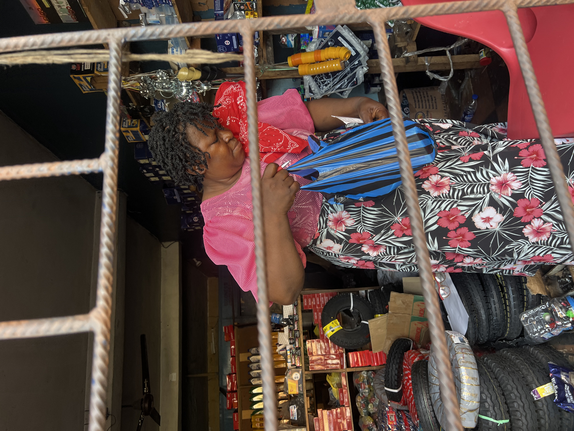 The image captures an indoor scene, likely a retail shop or stall, viewed through a grid of metal security bars in the foreground. The perspective looks into a well-stocked and somewhat cluttered space.
The central subject is a person, appearing to be a woman, resting or sleeping on a makeshift bed covered with a dark fabric adorned with a vibrant pattern of red and white hibiscus flowers and green leaves. She is lying on her back, eyes closed, with a peaceful expression. She wears a bright pink, mesh-textured short-sleeved top, and a red patterned cloth is draped over her chest and shoulders. She has dark, tightly curled hair. In her hands, she holds a blue and black striped, fan-like object.
The shop is filled with various goods, particularly vehicle parts and accessories. To the right, a significant stack of tires of different sizes is visible, some wrapped in protective plastic. Text like "ULTRAPOWER" can be seen on some tire sidewalls. Shelves line the walls, packed with boxes, smaller packaged items, and hardware components such as chains and yellow-handled tools. A red plastic chair is partly visible in the upper right. On the floor near the resting person, a small plastic bottle of water with "Aquafina" clearly printed on its label can be seen. The general lighting suggests an indoor setting, possibly during the day with some artificial light, or at dusk. The visible merchandise indicates the shop deals with automotive or mechanical supplies.