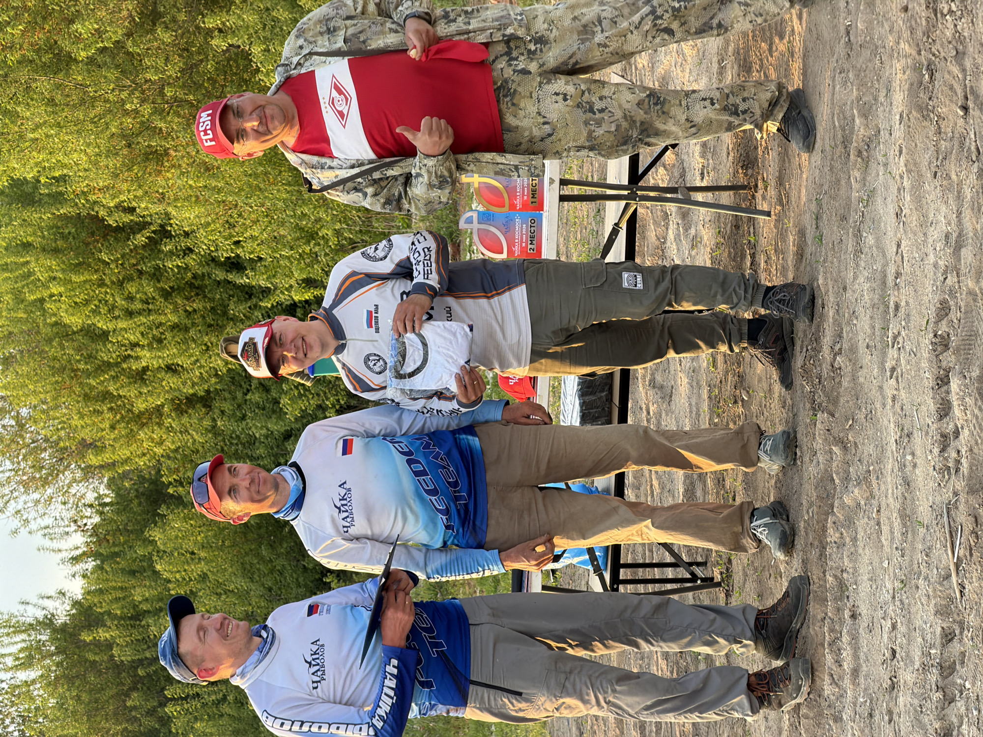 The image shows a group of four men standing outdoors on a dirt ground, possibly at a competition event. In the background, there's lush green foliage.
From top to bottom:
* The top man is wearing a red baseball cap with the letters "FCSM" and a red t-shirt with the logo of the FC Spartak Moscow soccer club, camouflage jacket, and giving a thumbs-up.
* The second man wears a white cap and white jacket, and is holding a white shirt. On a table behind, there are award trophies or plaques that read "ЧАЙКА В КОСМОСЕ", which translates to "Seagull in Space", and "1 MECT" and "2 MECTO", indicating 1st and 2nd places. The dates May 16, 2026, and May 16, 2020, are visible.
* The third man is wearing a blue and white jacket with the word "ЧАЙКА" and "FEEDER TEAM" on it. He wears a red cap.
* The bottom man is wearing a jacket with the word "ЧАЙКА" and "FEEDER TEAM" on it.
The awards indicate a connection to Krasnyi Bor and the "Seagull in Space" competition.