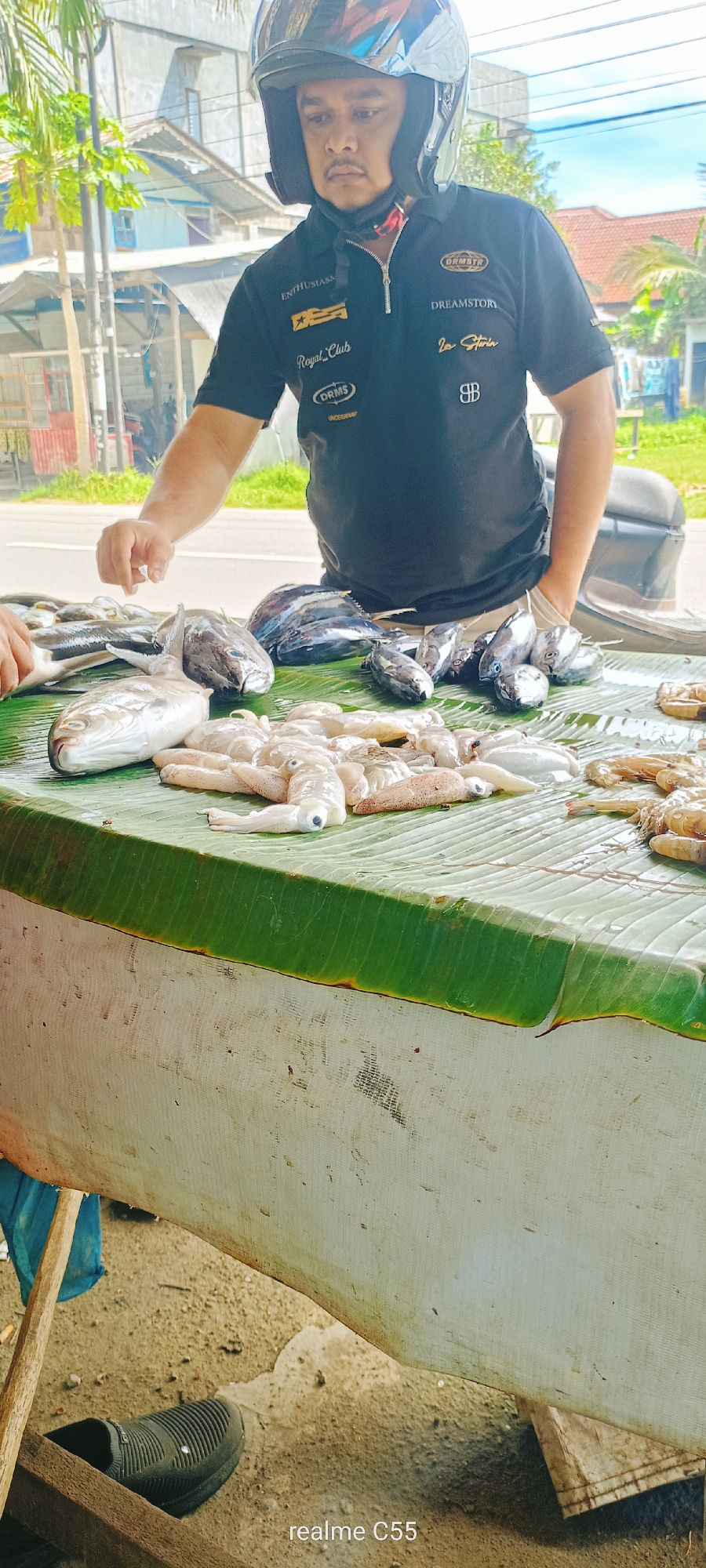 The image depicts an outdoor seafood market stall in Lhokseumawe, Indonesia. A male vendor, visible from the chest up, is positioned behind a display of fresh seafood. He wears a black polo shirt featuring various embroidered logos: "ENTHUSIASM," "Royal Club," "DRMS," and "UNDERGRAND" on the left side, and "DRMSTR," "DREAMSTORY," "Lee Storia," and a "BB" logo on the right side. A dark motorcycle helmet with a secured chin strap and a partially reflective visor covers his head. The man has a mustache and a neutral expression. His right hand is extended over the seafood, and a portion of another person's hand is visible on the far left, also near the seafood.
The seafood is arranged on green banana leaves, which cover a table. The display includes several whole fish of varying sizes (some appearing to be tuna or mackerel), a pile of white cephalopods (likely squid or cuttlefish) with visible eyes, and a collection of prawns or shrimp towards the right. Other fish are stacked in the background.
The table's sides are draped with a soiled, white, fine-mesh fabric or netting. Below the table, a wooden support structure and a blue fabric item are visible. The ground is a mix of bare dirt and concrete patches, with a black slip-on shoe present on the left side. The background shows a paved road, multiple buildings (appearing residential or commercial), and green tropical foliage under a bright sky. A "realme C55" watermark is located at the bottom center of the image.