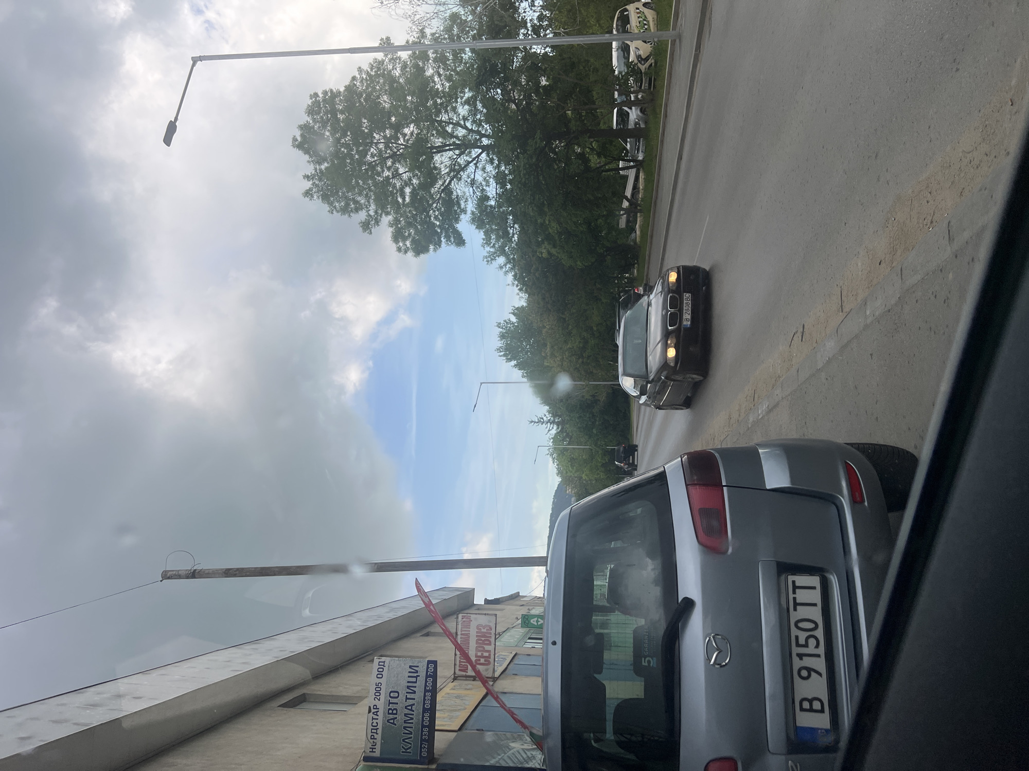 The image shows a street scene, likely in an urban area, based on the buildings and vehicles present. A silver Mazda is in the foreground, its Bulgarian license plate reading "B 9150 TT." A black BMW with a "B 2808 BC" plate is in front of the Mazda. The Mazda and BMW are in traffic on a paved road alongside a building. There are other vehicles parked or moving on the opposite side of the road. The building has signs on it, including one that reads "НОРДСТАР 2005 ООД АВТО КЛИМАТИЦИ 052/336 006; 0898 500 700" and another that says "АВТО СЕРВИЗ." Trees and some greenery are visible in the background, along with utility poles and streetlights. The sky is cloudy with patches of blue. Based on the license plates and the signs written in Bulgarian, this location is identified as Varna, Bulgaria.