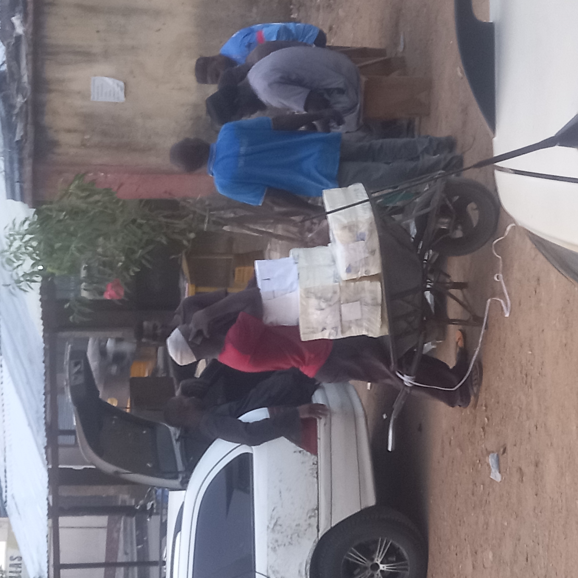 The image captures an outdoor scene in Dadi Riba, Nigeria, featuring several men, a pushcart, and a white car. The ground is unpaved and dusty.
**Subjects:** At least six men are visible in the frame. A black metal pushcart, or handcart, is prominently positioned, loaded with numerous stacks of packaged goods. A white sedan car is parked to the right, with its trunk open. Simple building structures and green foliage form the background.
**Activity or event taking place:** The men are gathered around the pushcart and the car, suggesting an active distribution or sale of goods. One man wearing a red shirt and a white cap is positioned near the front of the cart, while another in a blue shirt stands behind it. A third man in a dark shirt is seen interacting with the open trunk of the white car, either loading or unloading items. The overall impression is one of a bustling street-side exchange or commerce.
**Notable details:** The items on the pushcart are uniformly stacked, light-colored, rectangular packages, possibly loaves of bread or other food items, some wrapped in clear plastic. The lighting appears soft, indicating either early morning or late afternoon. The simple architecture of the background buildings and the dusty ground are consistent with an outdoor market or street setting in the specified location.
**Visible text:** A small, crumpled piece of paper is taped to the light-colored wall in the background, but no text on it is legible. A partial word, possibly "LAS" or similar, is faintly visible on a structure to the far left of the image.
