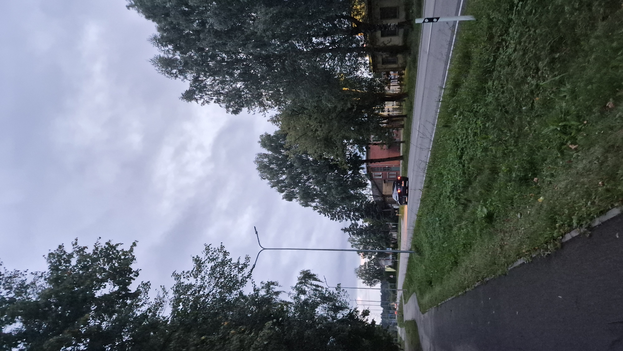 A low-angle, long shot captures a twilight scene on a road in Krauja, Latvia. The sky is overcast and gray, with scattered clouds. A paved road curves to the left, bordered by a grassy verge and trees on both sides. Tall, slender streetlights stand along the right side of the road. On the right side of the road, a dark-colored sedan is driving away from the camera, its headlights on. Buildings with red brick facades and lighter-colored structures are visible behind the trees on the right. A paved pedestrian path runs parallel to the road on the left side. The overall impression is of a quiet, somewhat overcast evening on a suburban or rural road.