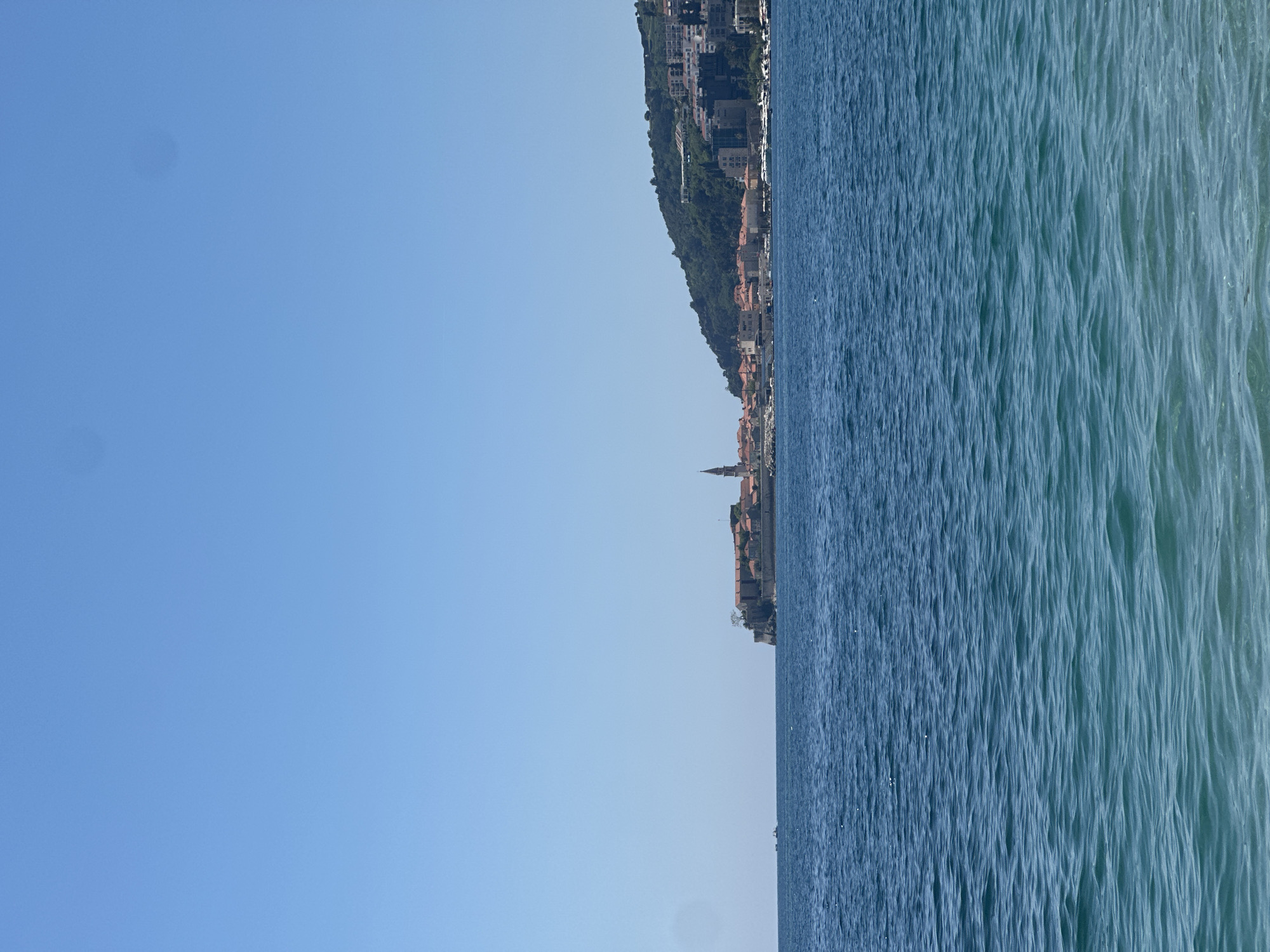 The image presents a serene coastal landscape, likely captured from the sea looking towards land. The scene is split horizontally into three main sections: a clear sky, the sea, and a town nestled against a green hillside along the horizon.
The upper portion of the image is filled with a vast expanse of clear, bright blue sky, indicating fair weather. Two faint, dark circular smudges are visible in the upper left section of the sky, consistent with dust spots on a camera lens or sensor.
The lower half of the image is dominated by the blue-green waters of the sea. The water's surface is textured with numerous small ripples and waves, catching the sunlight and creating varying shades of blue and turquoise. The sea extends from the foreground to the distant horizon.
Along the right side of the horizon, a verdant hill rises, densely covered in trees and other foliage. At the base of and extending partly up this hill, a coastal town or city is visible. The buildings feature traditional architecture, many with reddish-brown tiled roofs. A prominent, slender church steeple or tower, light in color with a conical top, stands out among the structures in the lower-right part of the town, suggesting a historical or central area.
The image depicts a peaceful daytime scene, under a bright sun, with no visible clouds. No people or specific activities are discernible. The setting, with its hilly coastline and characteristic architecture including a prominent steeple, strongly aligns with the provided context of Budva, Montenegro. There is no visible text in the image.