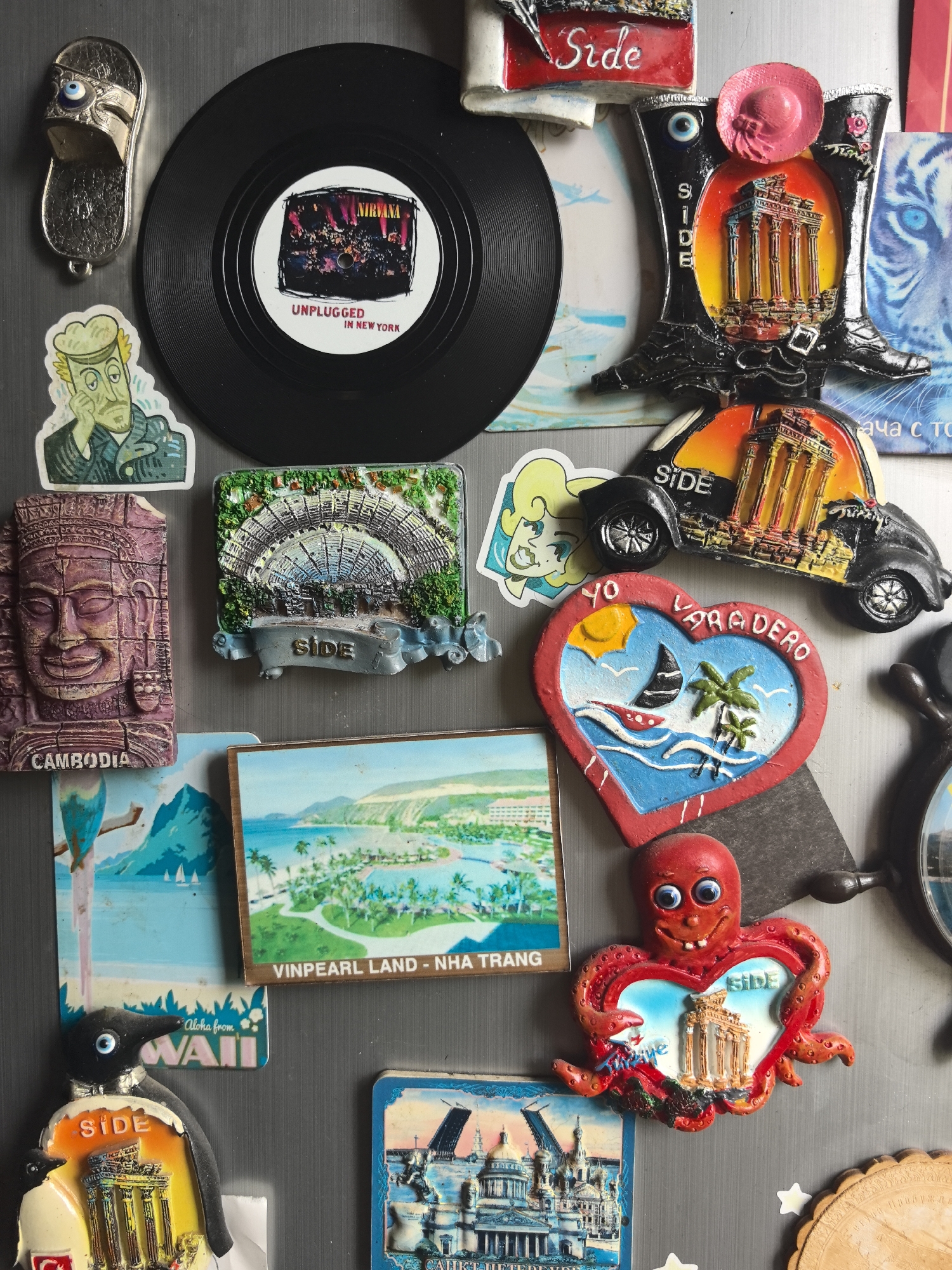 The image shows a refrigerator door covered in magnets and stickers. There is a vinyl record with the text "NIRVANA UNPLUGGED IN NEW YORK," along with various souvenir magnets from locations such as CAMBODIA, Varadero (Cuba), Hawaii ("Aloha from WAII"), and Nha Trang ("VINPEARL LAND - NHA TRANG"). A number of magnets feature the word "SIDE" alongside depictions of ancient Roman ruins. There are also decorative magnets like a sandal, a cartoon man, and an octopus. A sticker of a blonde woman is visible, as well as a magnet showing a tiger's face. No location of the city of Noviy, Russia is depicted within the image.