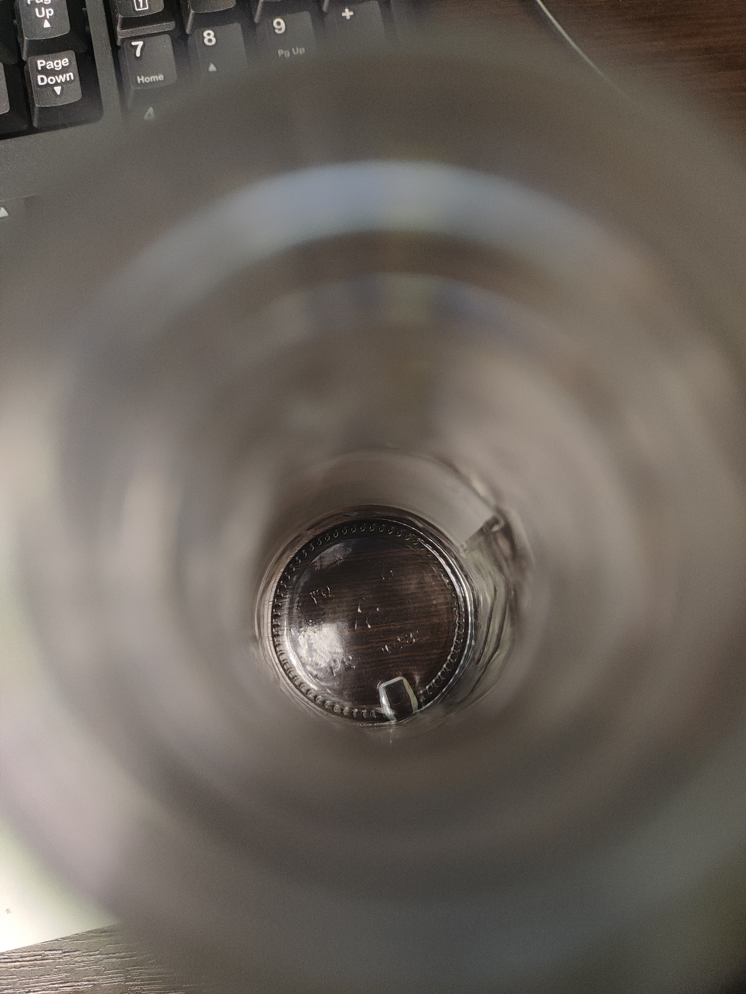 A close-up shot looking down into an empty clear glass bottle, showing the bottom of the bottle with a ridged edge. The bottle appears to be sitting on a wooden surface, which is also near a portion of a computer keyboard. The location is Unknown, Unknown.