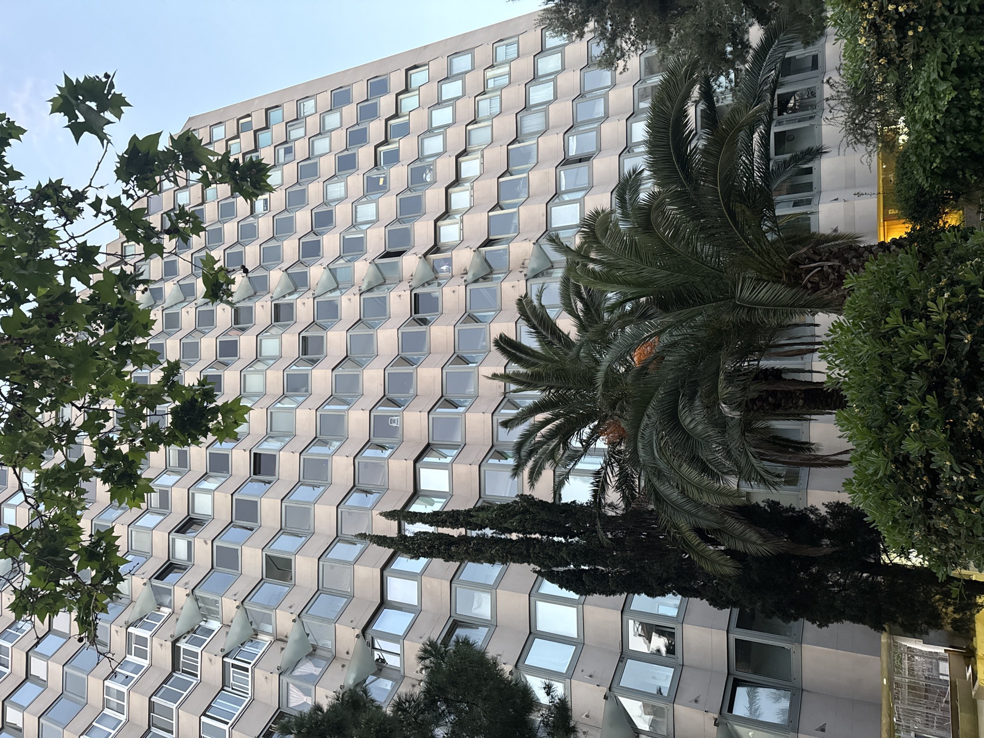 A low-angle, exterior shot of a modern building with a distinctive architectural style. The facade is characterized by repeating square windows that are set at an angle, creating a staggered, step-like pattern across the building's surface. Many of the windows are open, revealing glimpses into the interior. Various trees are in front of the building, including deciduous trees on the left and palm trees on the right. The sky above is pale and overcast. The scene is likely in Barcelona, Spain, given the architectural style and the presence of palm trees, which are commonly found in the city.