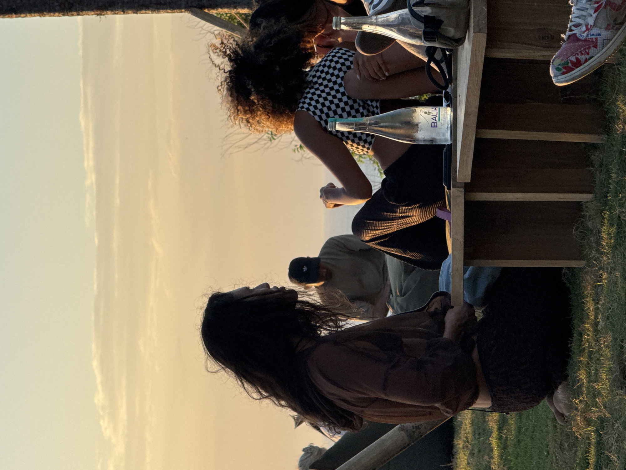 The image depicts a group of people gathered outdoors during what appears to be sunset, likely in Kuta Selatan, Indonesia, given the context provided. In the foreground, to the left, a person with long dark hair is seen from the side, wearing a loose, dark brown, sheer top and a dark textured skirt, kneeling on the grass. Facing them, to the right, is another person with curly dark hair, wearing a checkered crop top and dark pants, seated on a wooden bench. Between them, on the bench, are two glass bottles, one with a label that reads "BALIA". Behind these two individuals, further in the background, a person wearing a black baseball cap and a light-colored shirt is seated. The environment consists of green grass in the foreground and a bright, hazy sky with wispy clouds in the background, indicative of a sunset over what might be a coastal area. The overall mood is relaxed and informal.