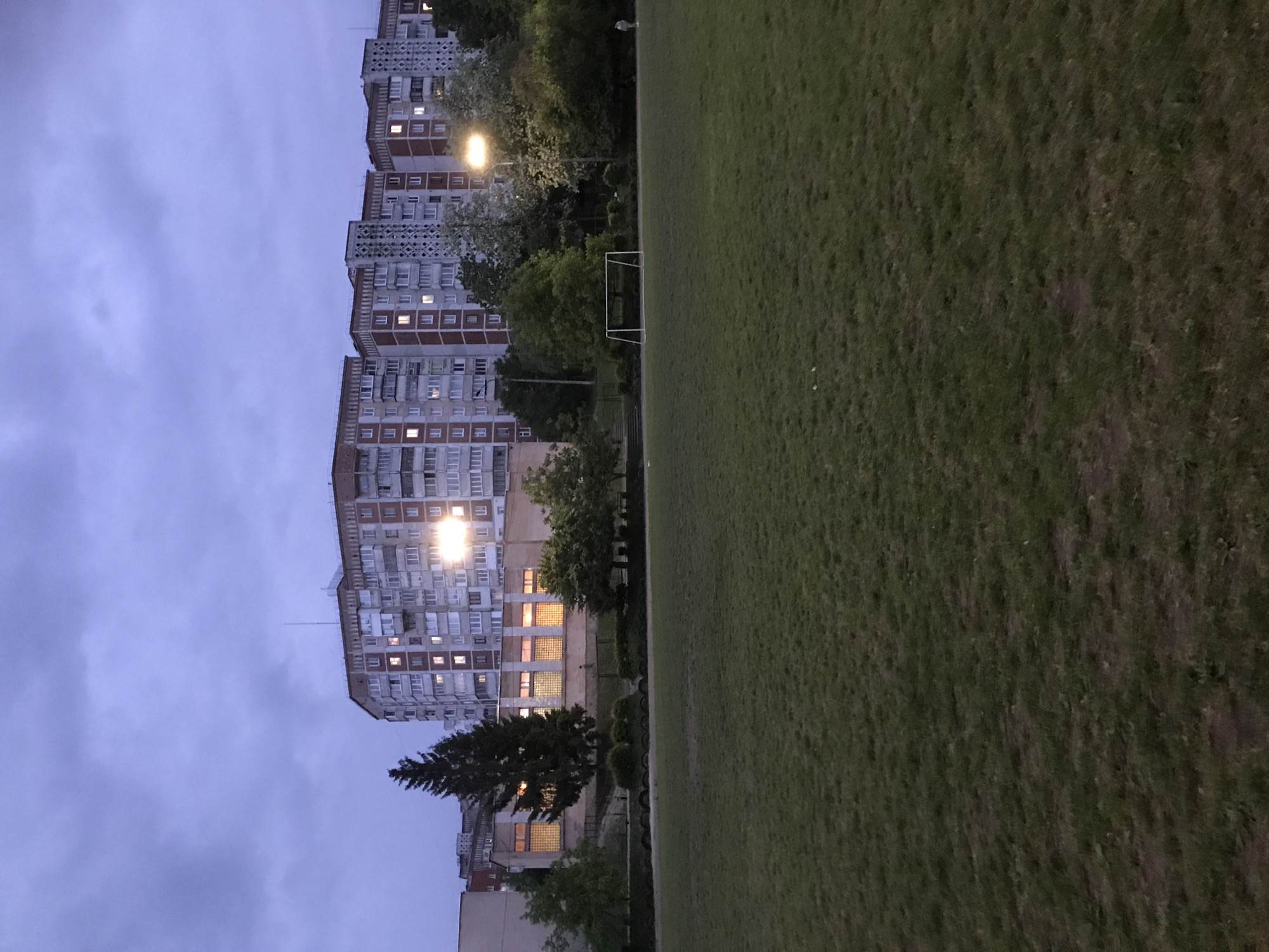 The image shows an outdoor scene at dusk, featuring a grassy field and a row of tall apartment buildings. The buildings are multi-storied and appear to be of Soviet-era construction, with repetitive window patterns. Some windows are illuminated, suggesting people are inside. A streetlight illuminates the area near the buildings. Between the buildings and the field, there are trees and some smaller structures. A soccer goal is visible on the edge of the field. The sky is overcast. The photograph is taken in Ternopil, Ukraine.