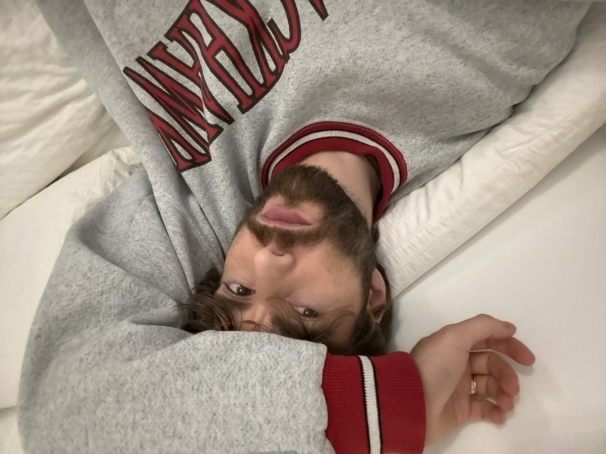 A person is lying on a white bed, wearing a gray sweater with red and white trim on the collar and cuffs and large red lettering across the chest. The person has light brown hair, a beard, and is looking directly at the camera. The photo is taken from an overhead perspective, with the person's head near the bottom of the frame and their body extending upwards. The environment suggests a bedroom setting, with white bedding. The location in the city of Montreal, Canada cannot be determined from the image.