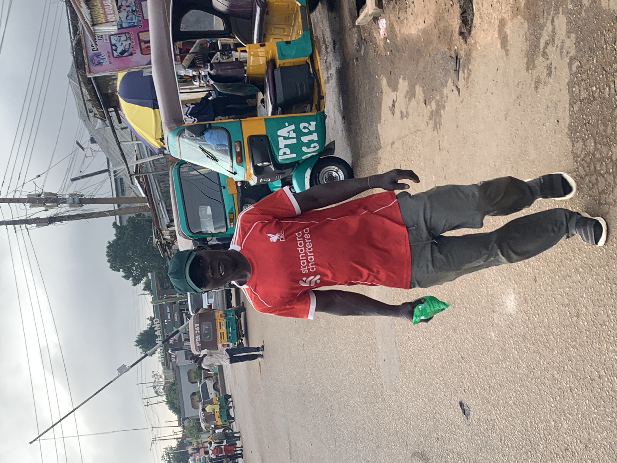 The image depicts a street scene in Jos, Nigeria, under an overcast sky. A man in the foreground, wearing a red Liverpool FC jersey and gray pants, walks towards the camera holding a crumpled green object in his left hand. He is also wearing a green baseball cap and black and white sneakers. Behind him, several auto-rickshaws, also known as "Keke Napep," are visible. One auto-rickshaw, painted yellow and teal, has "PTA-1612" written on its side. Further in the background, buildings with corrugated iron roofs and various signage are present, suggesting a commercial area or market. A white pole, likely part of a barrier or structure, extends diagonally across the upper left portion of the image. The road surface appears to be asphalt with some patches of dirt and debris.