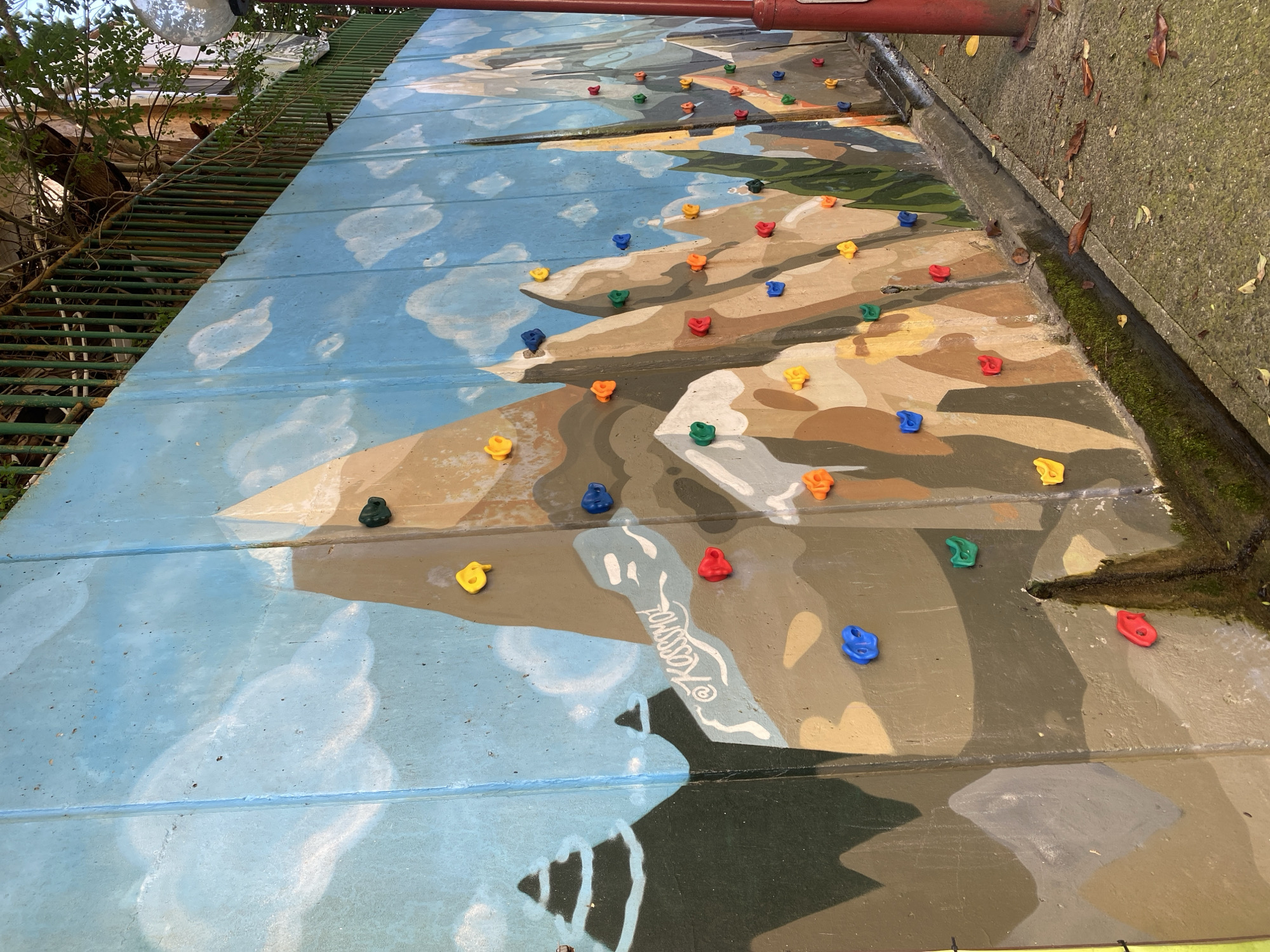The image shows a colorful mural painted on a concrete wall, depicting a mountain landscape with a bright blue sky and fluffy white clouds. Attached to the mural are numerous plastic climbing holds in various vibrant colors, including red, blue, yellow, orange, and green, indicating it is a climbing wall. The mural is positioned outdoors, adjacent to a concrete sidewalk or path and a narrow drainage channel. Overgrown vegetation and tree branches are visible at the top of the wall, and a red metal pole is present on the right side of the frame. Some fallen leaves are scattered on the ground. The mural features a signature that appears to read "@Kassmol". Based on a similar image, this location is identified as a climbing wall located in the city of Sochi, Russia.