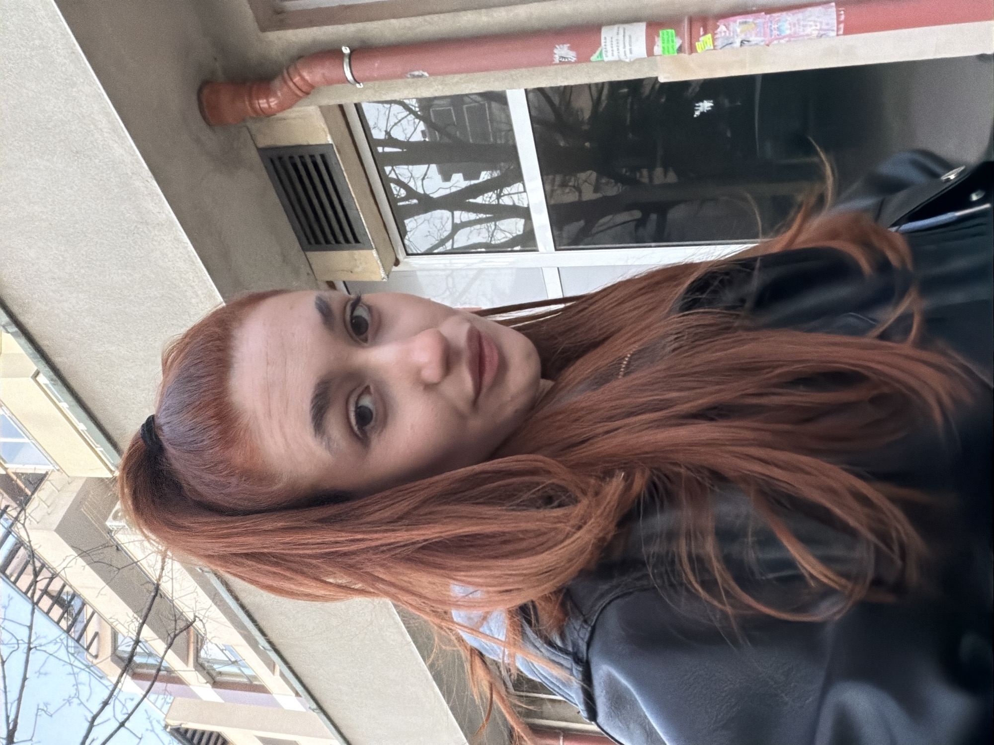 The image shows a woman with reddish-brown hair pulled back in a ponytail, wearing a black leather jacket. She is standing outside, and the background includes buildings with windows and tree branches without leaves. A red pipe and a window frame are visible in the upper portion of the image. Based on the architectural style of the buildings, the location is likely within the city of Plovdiv, Bulgaria.