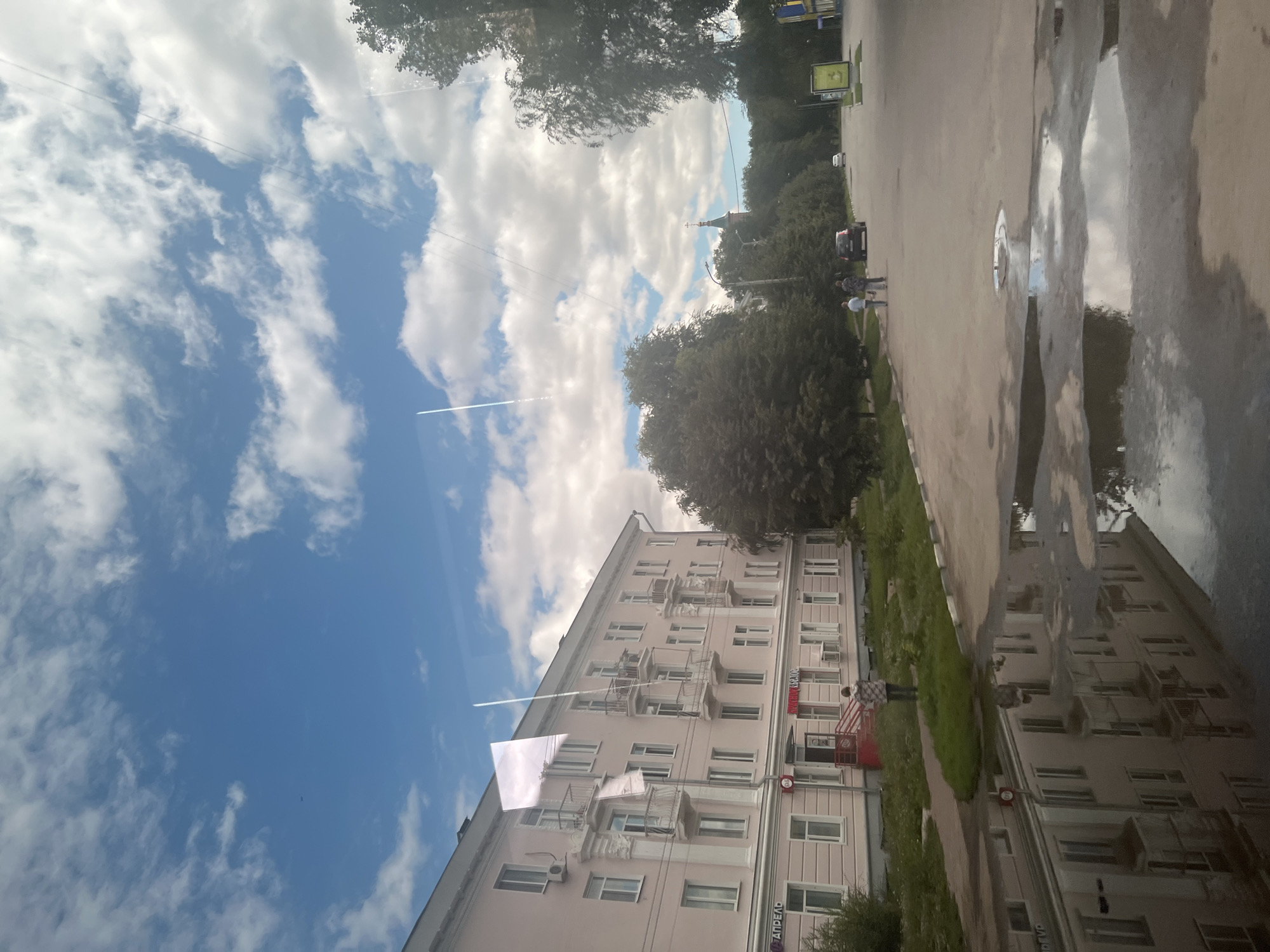 The image depicts a street scene in Ulyanovsk, Russia, with a large, light pink, multi-story apartment building on the left. The building has numerous windows, balconies, and air conditioning units. In front of the building is a sidewalk and a grassy area. A single person is standing on the sidewalk, facing away from the camera, in front of a red-carpeted staircase leading to an entrance.
The street in the foreground is wet, with large puddles reflecting the building and the sky. A few cars are visible on the road further down, along with trees and other buildings in the distance, including a church with a green dome and cross. The sky is blue with scattered white clouds, and contrails from aircraft are visible. Two people are walking on the sidewalk on the right side of the street, away from the apartment building. The overall impression is of a slightly overcast day in a residential area.