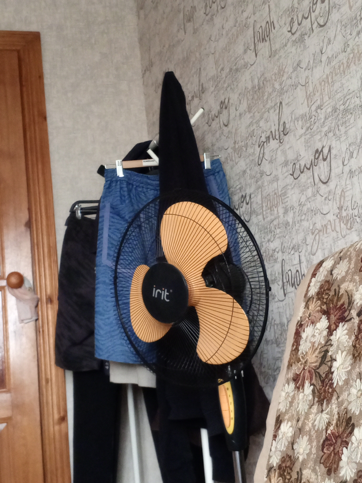The image shows an indoor scene in what appears to be a room. A black oscillating fan with orange blades and a black grill is positioned in the foreground, slightly off-center to the right. The fan has a black base and stem with a yellow accent. Behind the fan, clothing hangs on a rack, including a blue patterned garment and a dark garment. To the left of the fan and clothing, a wooden door with a round wooden knob is visible, along with a beige towel or cloth hanging on it. The wall behind the fan and clothing is covered in patterned wallpaper with cursive text. To the right of the fan, a portion of a floral-patterned fabric, possibly a blanket or upholstery, is visible. The lighting in the scene suggests it is daytime, with light coming from an unseen source. There are no people visible in the image.
The location is Naro-Fominsk, Russia.