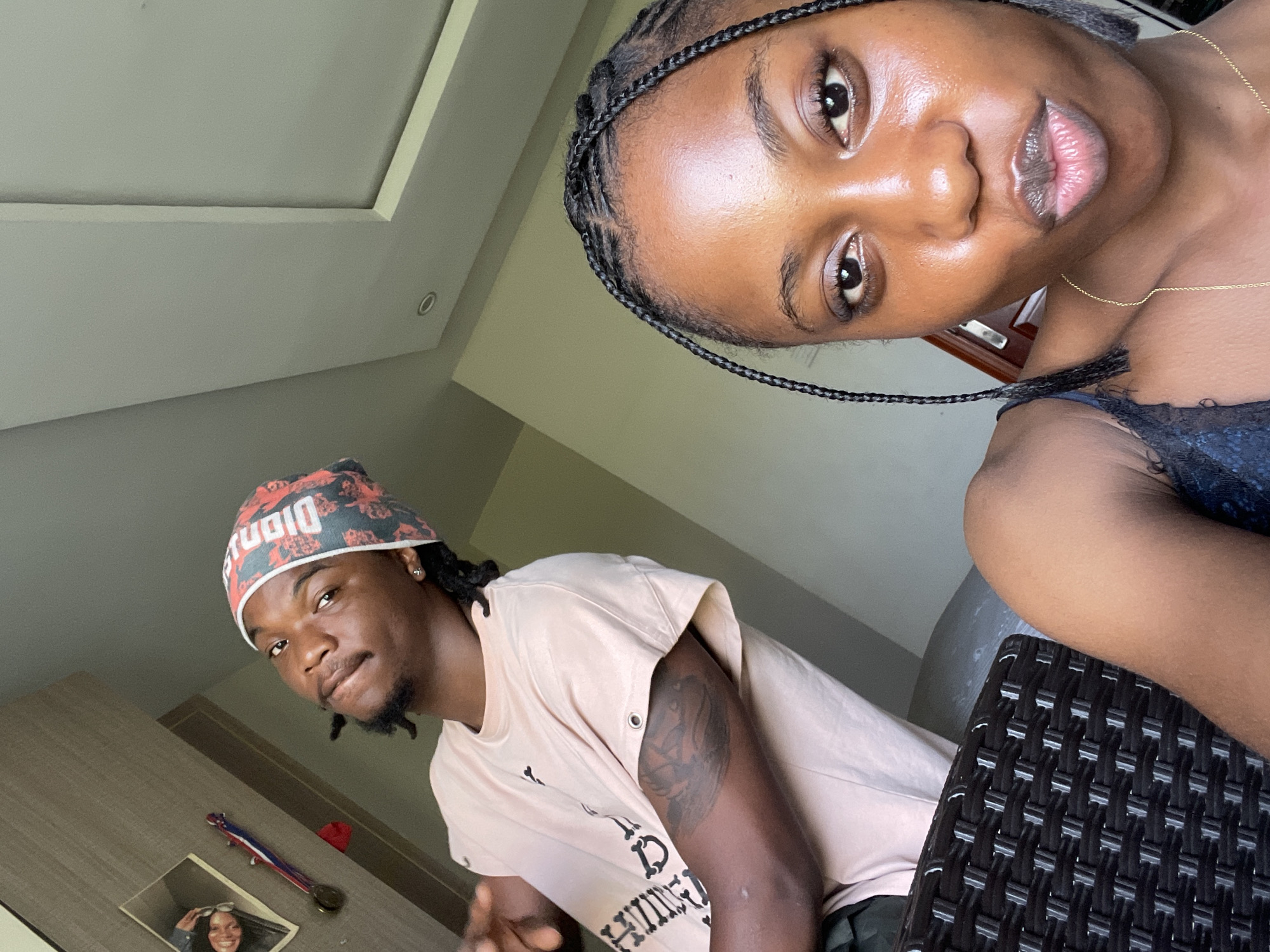 Two dark-skinned individuals are visible in an indoor shot. A woman with braided hair, positioned at the top right, looks directly at the camera. She wears a necklace and a dark-colored top. A man, wearing a bandana and a light pink t-shirt, is positioned on the left. He has a tattoo on his left arm. Behind the man, there is a cabinet with a framed picture on it. The background includes a ceiling and a window. The photo appears to have been taken in a private residence. The location is in the city of Unknown, Unknown.