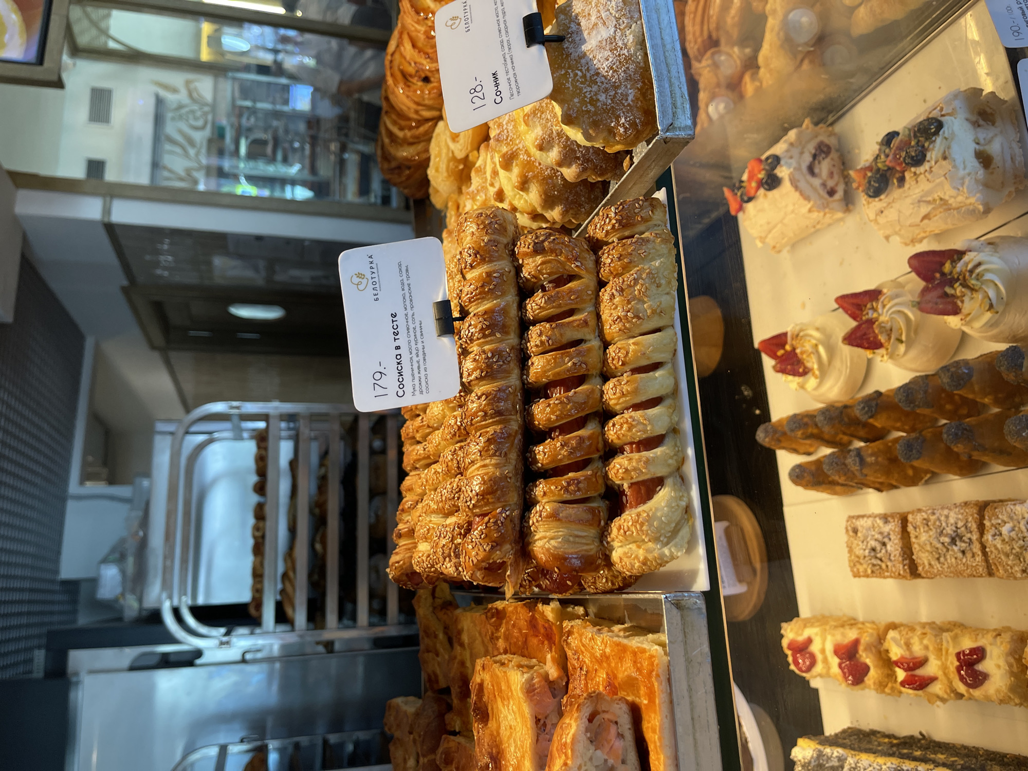 The image shows a display of baked goods in a bakery. In the foreground, there are several rows of pastries, including what appear to be sausage rolls wrapped in dough and sprinkled with sesame seeds. There are also some rectangular pastries with a dusting of powdered sugar, and small meringue-based desserts topped with berries and nuts. To the left, there are slices of a savory pie with visible filling, possibly salmon.
In the middle ground, behind the counter, shelves are filled with more baked goods, including braided loaves and other pastries. In the background, a person in a white uniform is partially visible behind a glass partition, seemingly working in the bakery. The overall environment is that of a retail food establishment, with display cases and shelving designed to showcase a variety of food items. The lighting suggests an indoor setting. There is a price tag visible for a "Сосиска в тесте" (sausage in dough) at 179, and another for "Сочник" (a type of pastry) at 128.