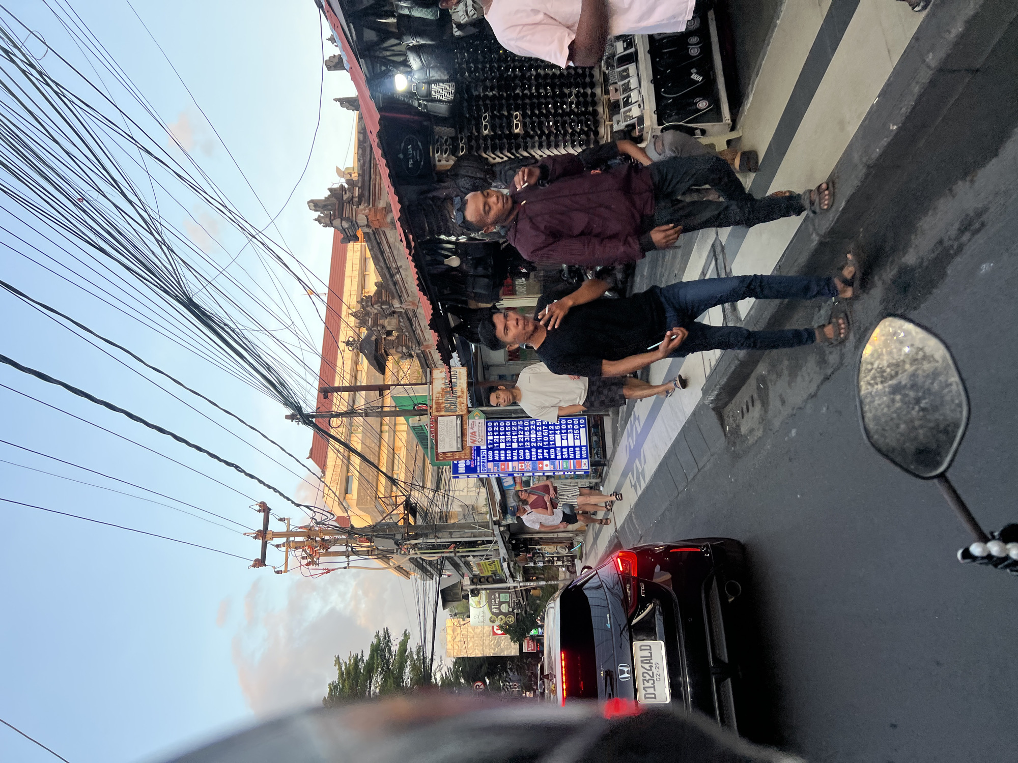 The image depicts a street scene in Kuta, Indonesia, likely during late afternoon or early evening, with golden hour lighting. In the foreground, a motorcycle's side mirror is visible, reflecting the asphalt road and some of the scene. A black Honda car, with its rear visible and license plate "D1324ALD", is positioned on the left side of the road.
The street is lined with shops and stalls. One stall displays various leather bags and sunglasses. Another stall features a currency exchange board with rates for USD, AUD, EUR, GBP, JPY, NZD, CAD, SGD, MYR, THB, and IDR. Several people are walking on the sidewalk and crossing the street.
On the right side of the image, three men are walking. The man in the center is wearing a maroon jacket and jeans, and is smoking a cigarette. To his left, a man in a black t-shirt and jeans is also smoking. To the far right, another man in a light-colored t-shirt and shorts is walking. Further down the sidewalk, a couple is walking, with the woman in a striped skirt and the man in a light-colored shirt and shorts.
Overhead, a complex network of electrical wires and poles dominates the sky, indicating a densely populated urban area. Buildings with traditional Balinese architectural elements are visible behind the stalls. The overall atmosphere suggests a bustling commercial area with pedestrian and vehicle traffic.