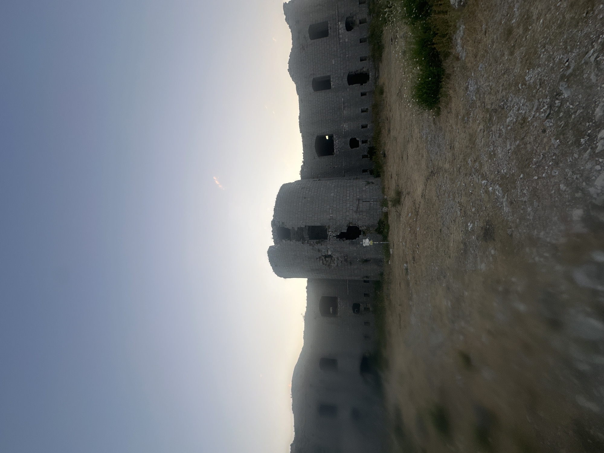 The image captures the ruins of the Brajici Fortress, located in Brajici, Montenegro. The fortress is constructed of stone blocks and shows significant signs of decay, with many sections of the walls missing, exposing the interior. Openings that were once windows or doorways are now dark voids. The architecture suggests a historical military structure with a prominent circular tower on the right side of the frame.
The foreground consists of dry, overgrown ground with patches of sparse vegetation and small white flowers. The terrain appears uneven and rocky. In the background, a hazy, mountainous landscape is visible under a pale sky that transitions from a light blue at the top to a softer, warmer hue near the horizon. A faint, thin cloud can be seen in the upper right portion of the sky. There are no people visible in the frame. The overall atmosphere is one of historical abandonment and natural overgrowth.
