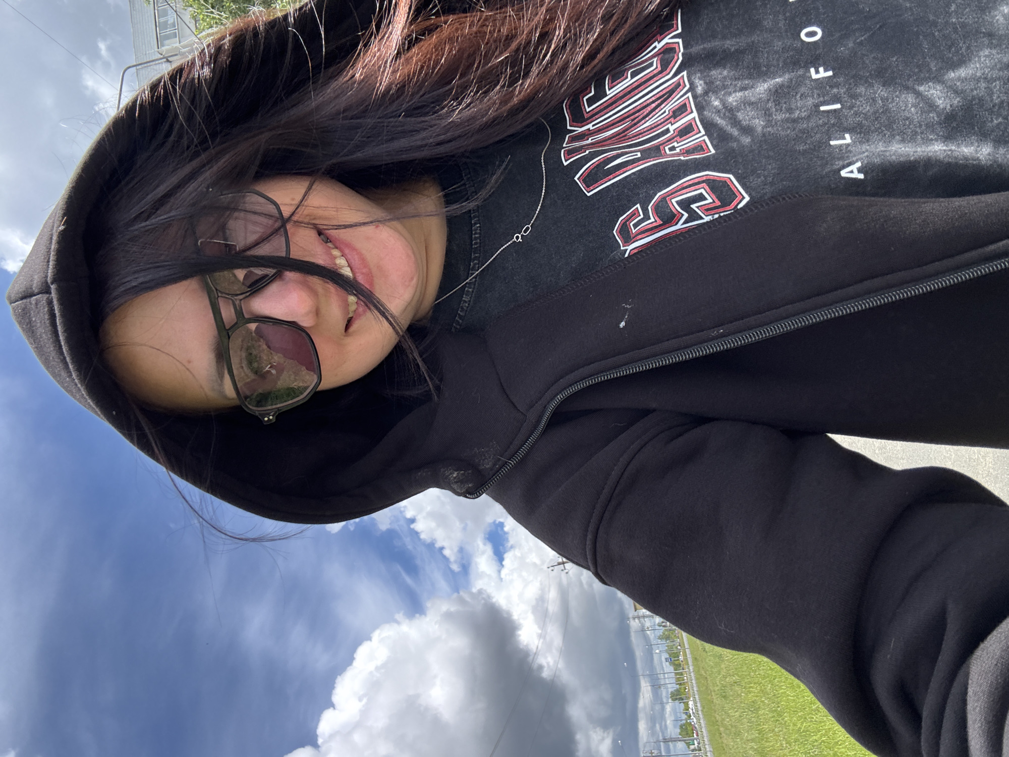 The image displays a person, likely female, captured in a rotated, upward-angled shot, facing the camera with a slight smile. The person has long, dark hair and is wearing dark-rimmed eyeglasses or sunglasses, which reflect the cloudy sky.
They are dressed in a black hooded sweatshirt or jacket, worn over a dark t-shirt. The t-shirt features prominent stylized text in white with red outlines. Clearly visible is the word "CALIFORNIA" oriented vertically, and above it, partially obscured letters that appear to spell "SAN", suggesting a university or location name like "SAN DIEGO". A thin silver necklace with a unique two-loop pendant is also visible around their neck.
The background shows an expansive, bright blue sky with numerous large, fluffy white and grey clouds. To the right of the subject, a vibrant green grassy field stretches into the distance. Faint structural elements, possibly power lines or distant buildings, and what appears to be a highway with vehicles are visible further back. The scene is set outdoors during the daytime, under bright, possibly partly cloudy, weather conditions. The overall impression is one of a casual, cheerful moment captured outdoors.