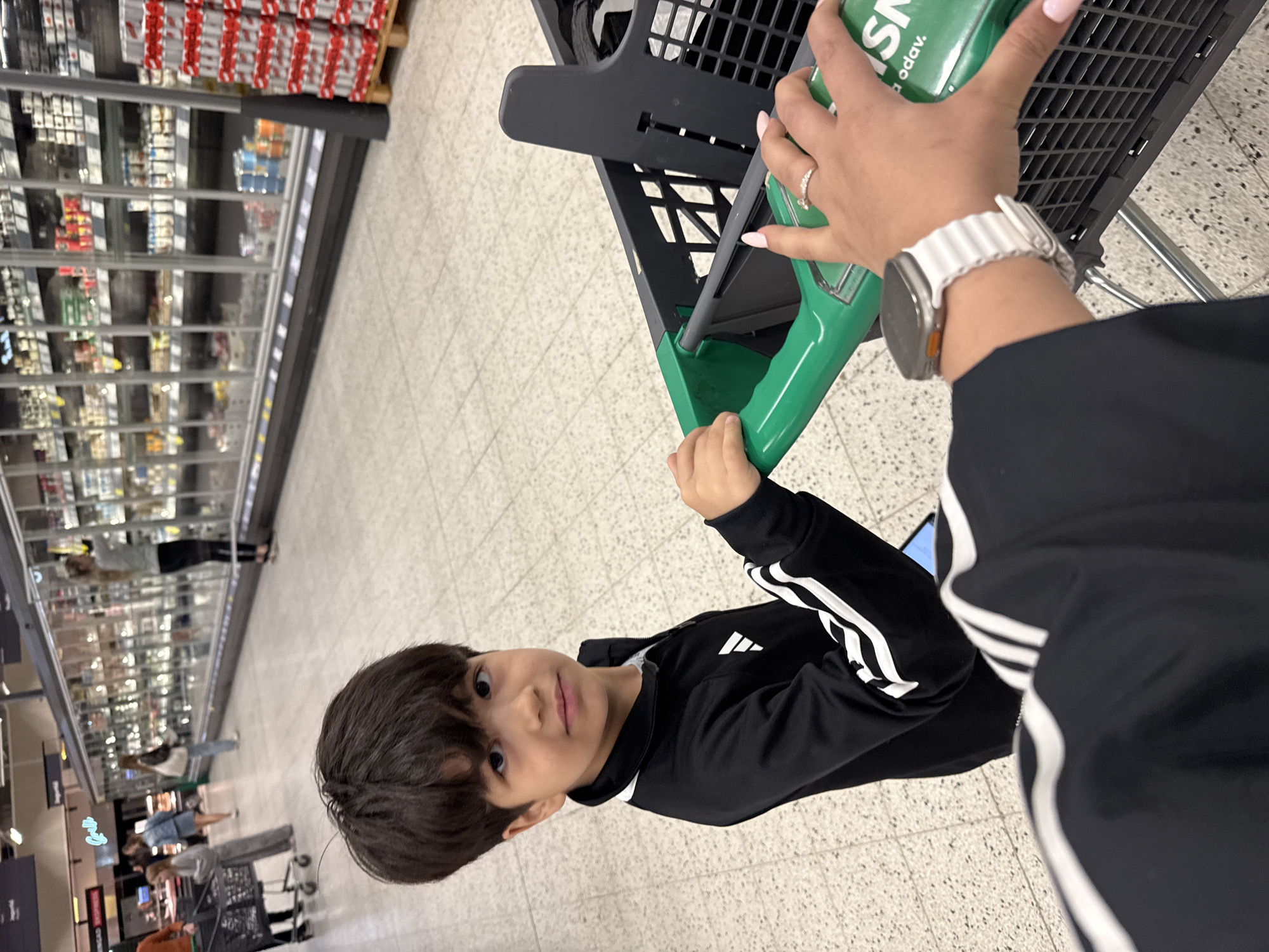 The image shows a young boy in the foreground of a supermarket in Tallinn, Estonia. He is wearing a black Adidas jacket with white stripes on the sleeves. The boy is looking towards the camera with a neutral expression. Behind him, a hand is visible pushing a shopping cart. The shopping cart has a green handle and a black basket. In the background, there are aisles of refrigerated display cases filled with products. Other shoppers can be seen in the distance, browsing the shelves or walking through the store. The floor of the supermarket is tiled with a light-colored pattern. A stack of red and white cans is visible on the right side of the image.