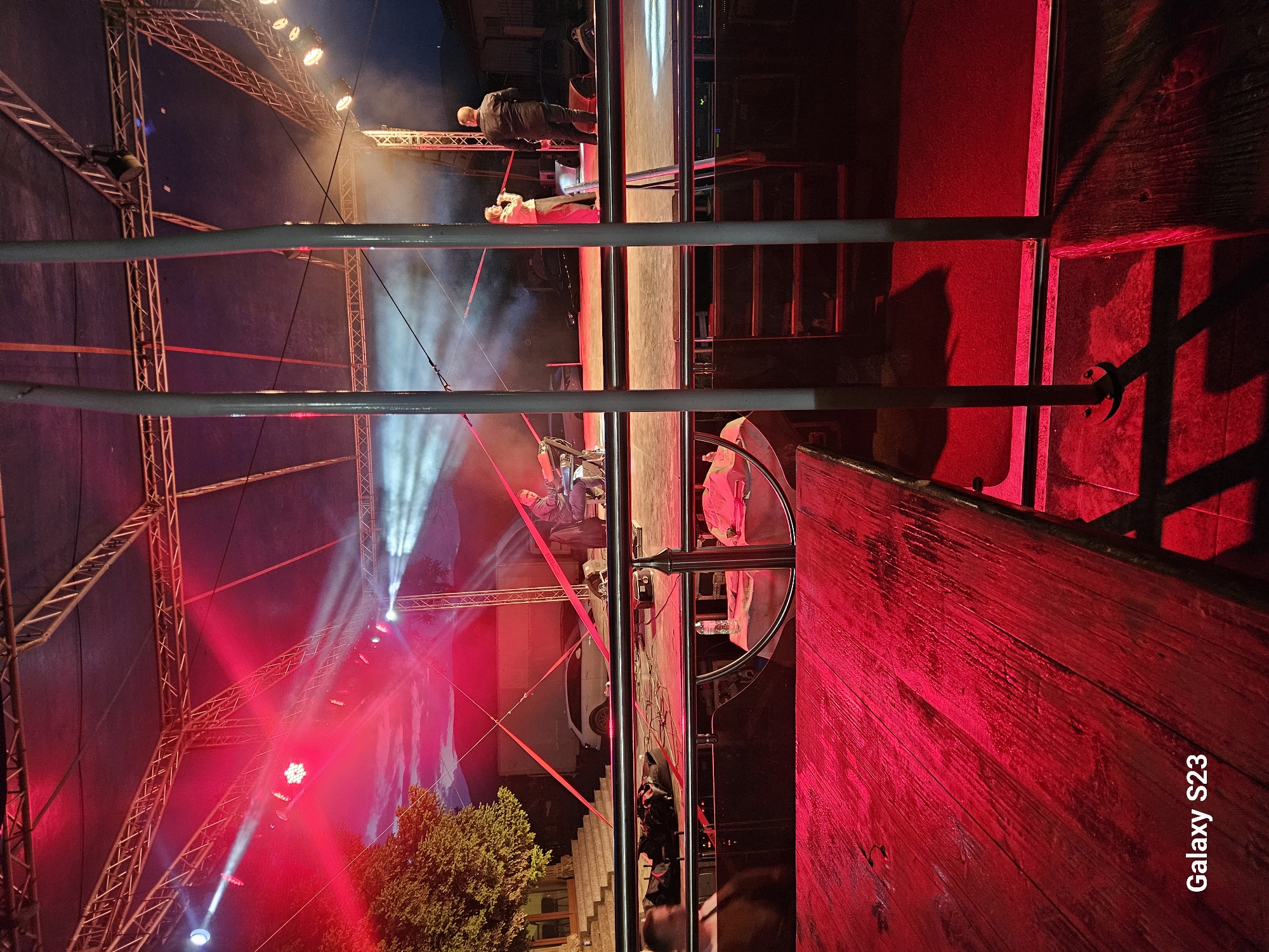 The image, apparently taken in Sliven, Bulgaria, shows an outdoor event, possibly a performance, taking place under a large tent-like structure illuminated with red and white stage lights. The camera viewpoint is partially obstructed by silver metal railing. In the background, figures are visible on a raised stage. The stage is lit with bright red lights, creating a dramatic effect. In the foreground, a wooden surface is prominently displayed, bathed in red light. The image appears to have been rotated 90 degrees counter-clockwise. A "Galaxy S23" watermark is visible on the bottom right corner.