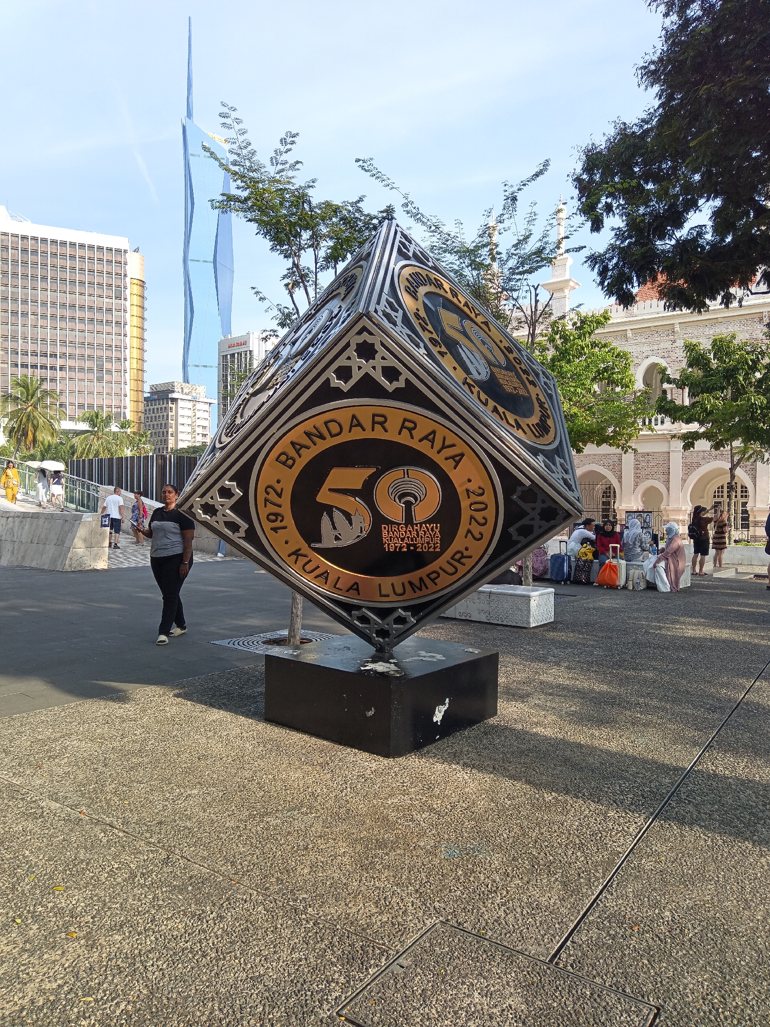 In Kuala Lumpur, Malaysia, a large, metallic sculpture is prominently displayed on a paved area. The sculpture, which appears to be a stylized cube or diamond, is adorned with intricate designs and text. One visible face of the sculpture prominently features the number "50" in gold, surrounded by circular text that reads "1972. BANDAR RAYA KUALA LUMPUR 2022". Below this, smaller text indicates "DIRGAHAYU BANDAR RAYA KUALA LUMPUR 1972-2022".
In the background, several modern buildings rise into a clear sky, including a tall, sleek skyscraper with a pointed top. To the right, an older, colonial-style building with arches and brickwork is visible, suggesting a mix of architectural styles in the area.
Several people are present in the scene. A woman in a grey and black t-shirt and dark pants walks across the paved area in the foreground. Further back, a group of people, some with luggage, are seated and standing near the older building, appearing to be waiting. Other individuals are seen walking in the background, suggesting a public urban space. The ground is paved with a coarse, textured material, and shadows from the sculpture and trees are cast across it. Lush green trees are scattered throughout the scene, adding natural elements to the urban landscape.