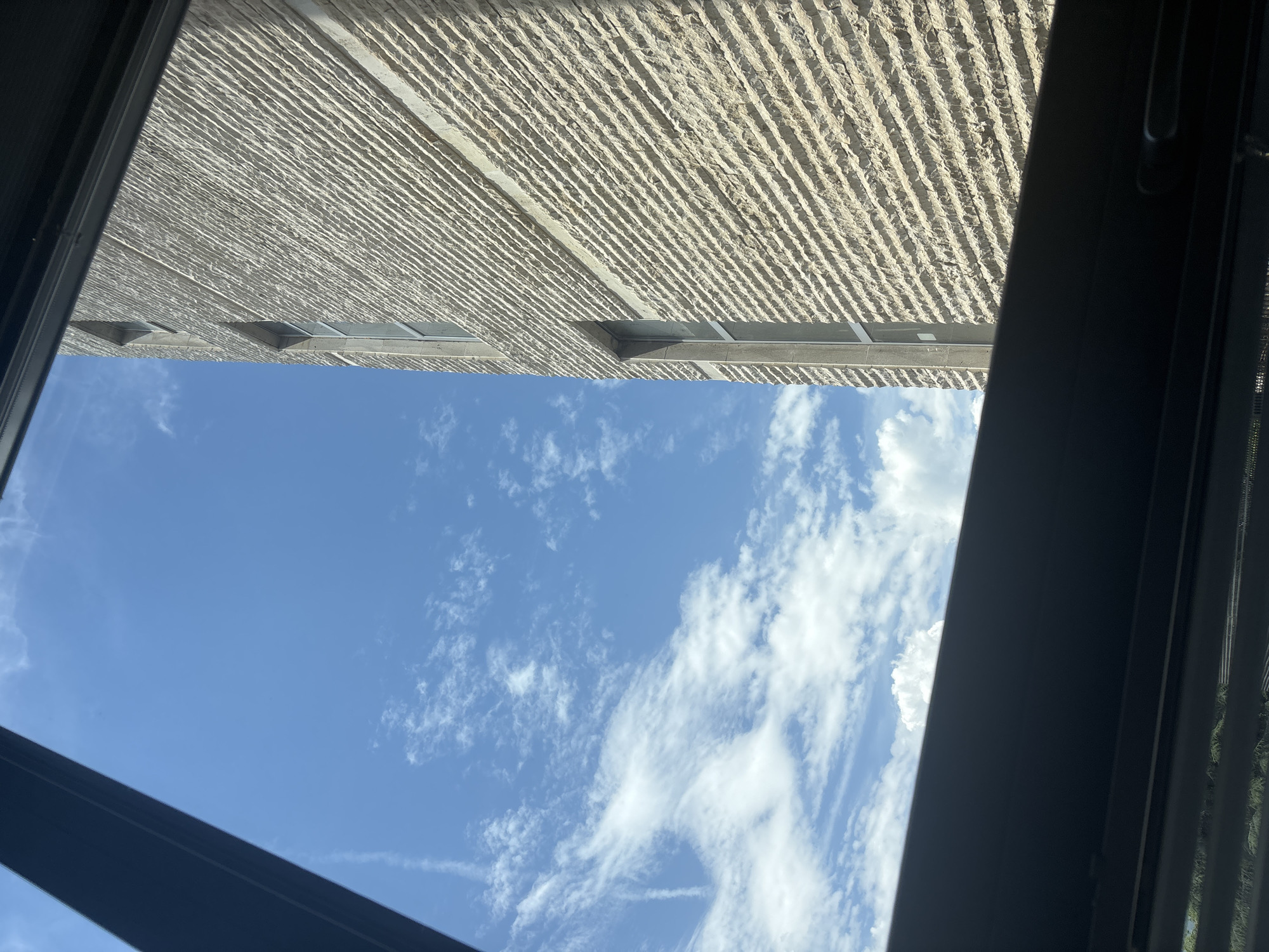 A vertically oriented photograph captures a view of a building and the sky, seemingly taken from an interior space looking upwards through a window or skylight.
The left side of the frame is dominated by a clear, bright blue sky filled with scattered white clouds. The clouds vary from wispy, high-altitude streaks to more defined, puffy cumulus formations, indicating fair weather.
The right side of the frame features a large, light-colored building. Its facade is distinctly textured with numerous horizontal grooves, giving it a stratified or ribbed appearance. Several rectangular recesses or windows are visible along the building's side, interrupting the textured surface.
Dark, possibly metallic or plastic, window frame elements partially obscure the view along the top and bottom edges of the image. A more substantial dark, diagonal beam runs across the lower left portion of the sky, further framing the view.
The scene depicts a sunny daytime. There are no people or discernible activities taking place. No visible text is present in the image. The architectural style of the building, with its textured facade, is a notable detail within the provided location context of Blagoevgrad, Bulgaria.