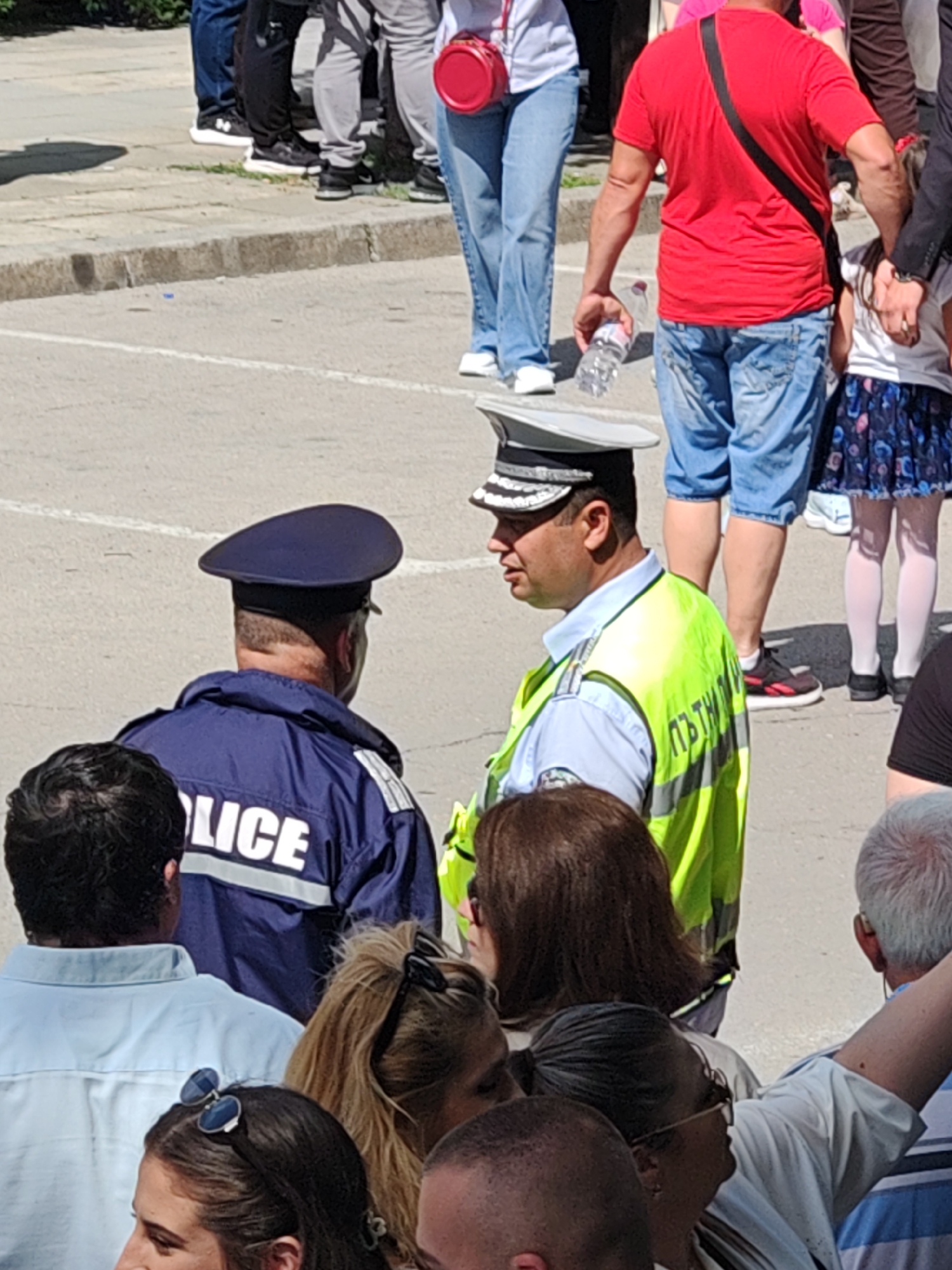 The image depicts a gathering of people in Shumen, Bulgaria. Two police officers are centrally positioned, one wearing a dark blue uniform with "POLICE" on the back, and the other wearing a white hat and a neon yellow vest over a blue shirt. Both are surrounded by civilians. A person wearing a red shirt and denim shorts holds a water bottle. Other individuals in the background are wearing various clothing styles. The environment appears to be an open outdoor space with concrete pavement, suggesting a street or public square.