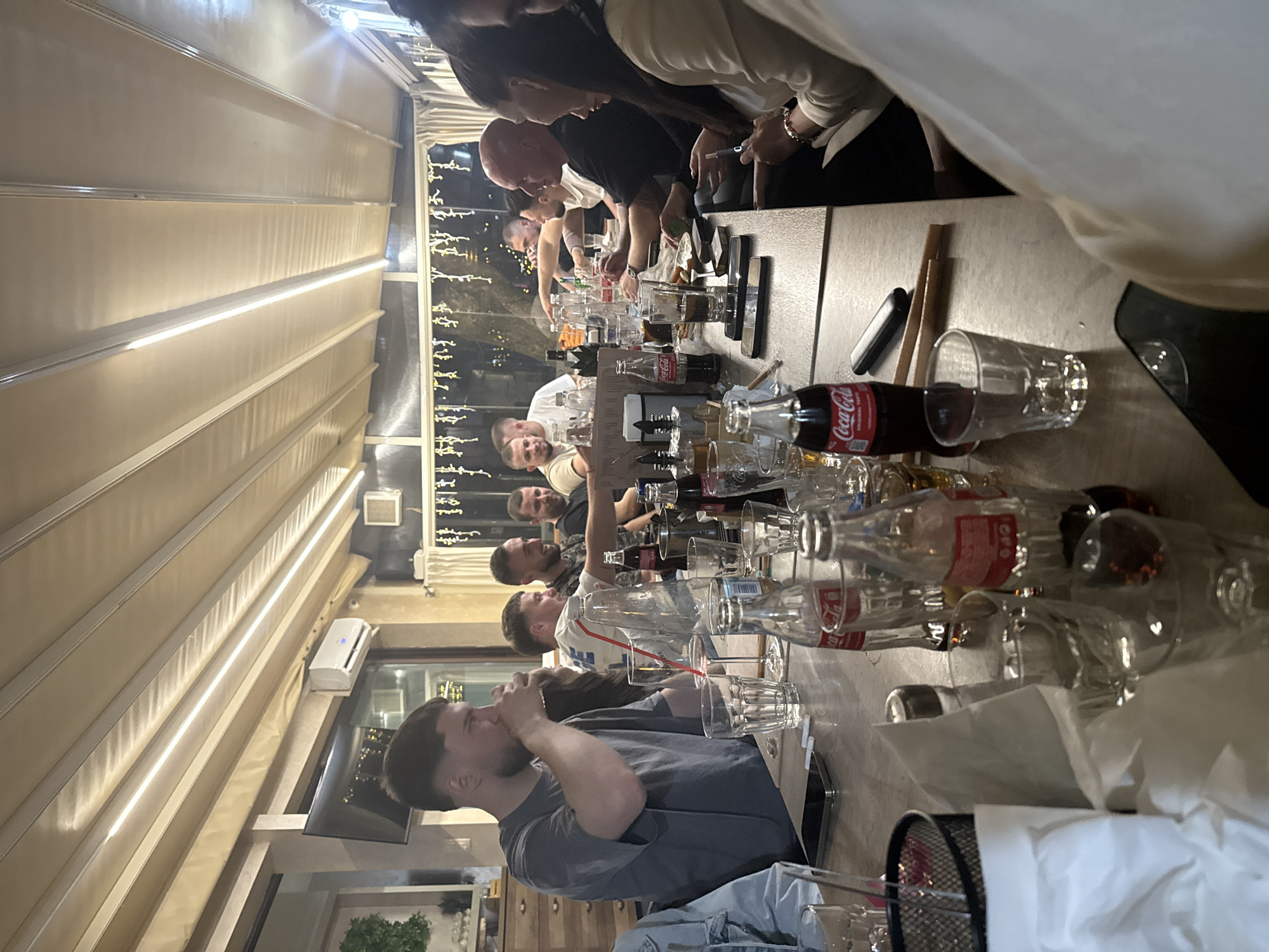 A group of people are gathered around a long table, appearing to be at a restaurant or outdoor eating area. The scene is brightly lit, with a canopy structure overhead. The table is cluttered with numerous empty Coca-Cola bottles and drinking glasses, indicating that a meal or gathering has recently occurred. Several individuals are visible, including one man in the foreground wearing a gray t-shirt and drinking from a glass. Other people are visible further down the table, engaging in conversation or other interactions. There are lights strung up in the background suggesting a festive or celebratory atmosphere.