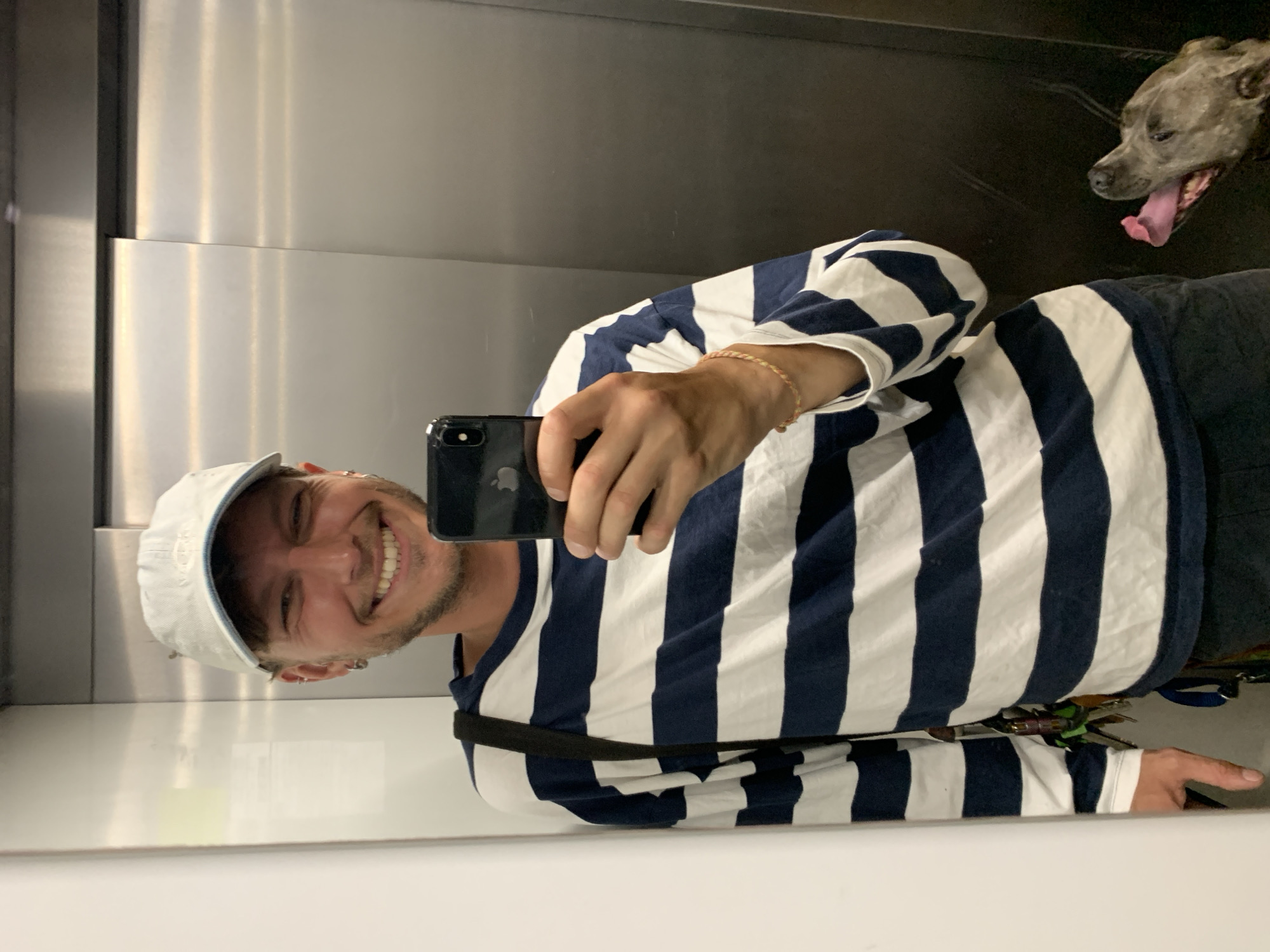 A selfie taken in an elevator with a man and his dog.
The man is fair-skinned, bearded, and wearing a white baseball cap, and a white and navy blue striped shirt. He's holding a black smartphone in his right hand, taking the picture. He is smiling. He wears multiple piercings in his left ear. He is wearing a thin braided bracelet on his right wrist.
The dog, which appears to be a grey-brown brindle pitbull, is panting with its tongue hanging out.
The background is the interior of a stainless steel elevator. The lighting appears to be fluorescent. A black strap is across the man's body. Keys and a lanyard are visible under his shirt near his right hip.
The image does not provide enough detail to infer a location in Tel Aviv, Israel.