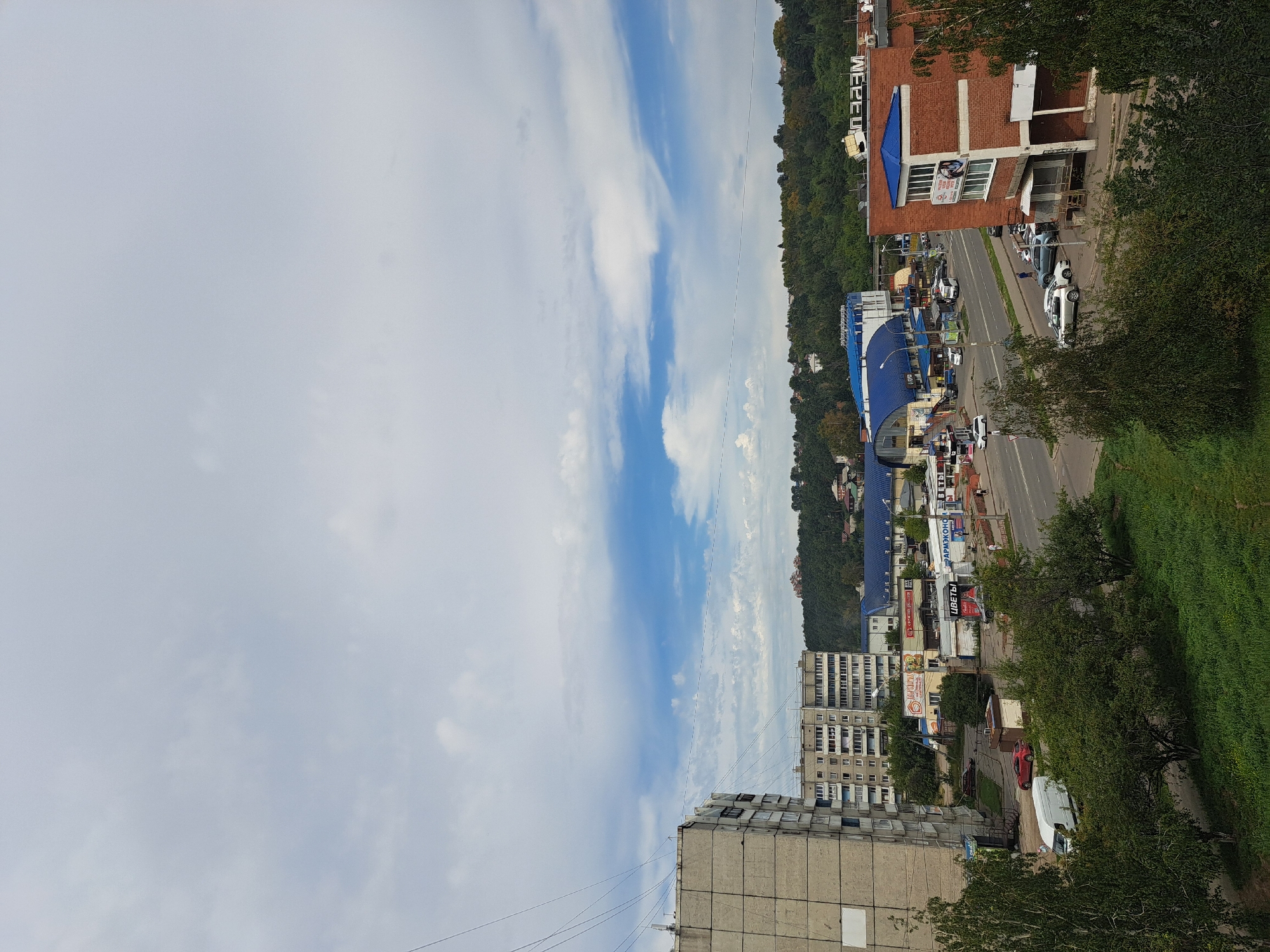 This is a medium-distance, eye-level shot of a street scene in Irkutsk, Russia, on a cloudy day. The foreground features a patch of green grass and several trees, with branches reaching into the frame. Behind the greenery, a two-lane asphalt road runs horizontally across the image, with vehicles and buildings lining either side.
On the left side of the road, several cars, including a white van, are parked or driving. A large, grey, multi-story apartment building dominates the far left of the frame, with visible windows and balconies. Adjacent to it are other apartment buildings with lighter facades. Several shops are visible along the street, some with blue awnings and signage in Cyrillic script, including what appears to be a pharmacy and a flower shop.
On the right side of the road, a modern brick building with a blue roof stands out, featuring multiple windows and a visible sign with Cyrillic text. This building has an elevated entrance and a small paved area in front. More trees and greenery are visible further back, obscuring the base of a densely wooded hill that forms the backdrop of the scene. The sky above is filled with scattered clouds, with patches of blue sky visible between them.
