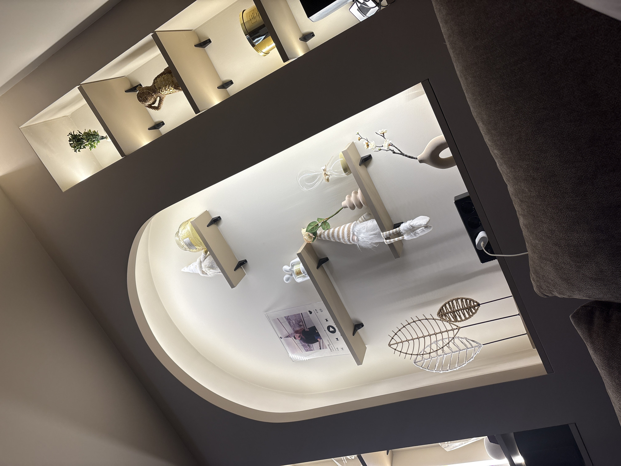 This image depicts an interior residential or commercial space, likely a living area, focusing on a built-in decorative wall unit. There are no discernible elements within the image to confirm a location in Sofia, Bulgaria.
The wall unit is composed of two primary sections: a large, deep arched recess on the left and a vertical stack of three rectangular, recessed niches on the right. Both sections feature integrated warm-toned LED strip lighting that illuminates their interiors. The wall surfaces within the unit are a light off-white or beige, contrasting with the darker grey-brown color of the unit's outer frame. The bottom right foreground shows a blurred, dark brown textured fabric surface, consistent with upholstery from a sofa, indicating the observer's low vantage point.
Within the arched recess, three light-colored shelves are supported by dark, L-shaped brackets. The topmost shelf holds a white, conical gnome figure with a striped cap and a translucent yellow glass object. The middle shelf displays a clear acrylic plaque featuring a photograph of two individuals, overlaid with a music player interface partially showing the song title "Falling" and artist "Trevor Daniel," alongside a small, clear glass sculpture resembling two stylized figures. The bottom shelf holds another white, conical gnome figure, a single light pink rose with a green stem in a small cylindrical, light-colored vase, a glass hourglass containing light-colored sand, and a small decorative branch with white blossoms in a C-shaped light-colored vase. On the surface directly below the bottom shelf of the arch, three stylized leaf sculptures made of woven white and brown materials stand on dark bases. Adjacent to these are a partially visible white spherical electronic device, a dark power strip with multiple outlets, a white power adapter plugged into it, and a small, dark-colored lamp with a flexible neck positioned to illuminate the display items.
The right section's three rectangular niches also contain light-colored shelves supported by dark L-shaped brackets. The top niche's shelf holds a small artificial green plant in a white pot. The middle niche's shelf displays a textured, light-brown sculpture, possibly an abstract figure, made of woven material. The bottom niche's shelf features a round, golden-colored tin with dark accents, partially labeled "COFFRE," and a segment of a dark electronic screen, possibly a television or monitor. No people are present in the immediate scene; however, a photograph of two individuals is depicted on an acrylic plaque.