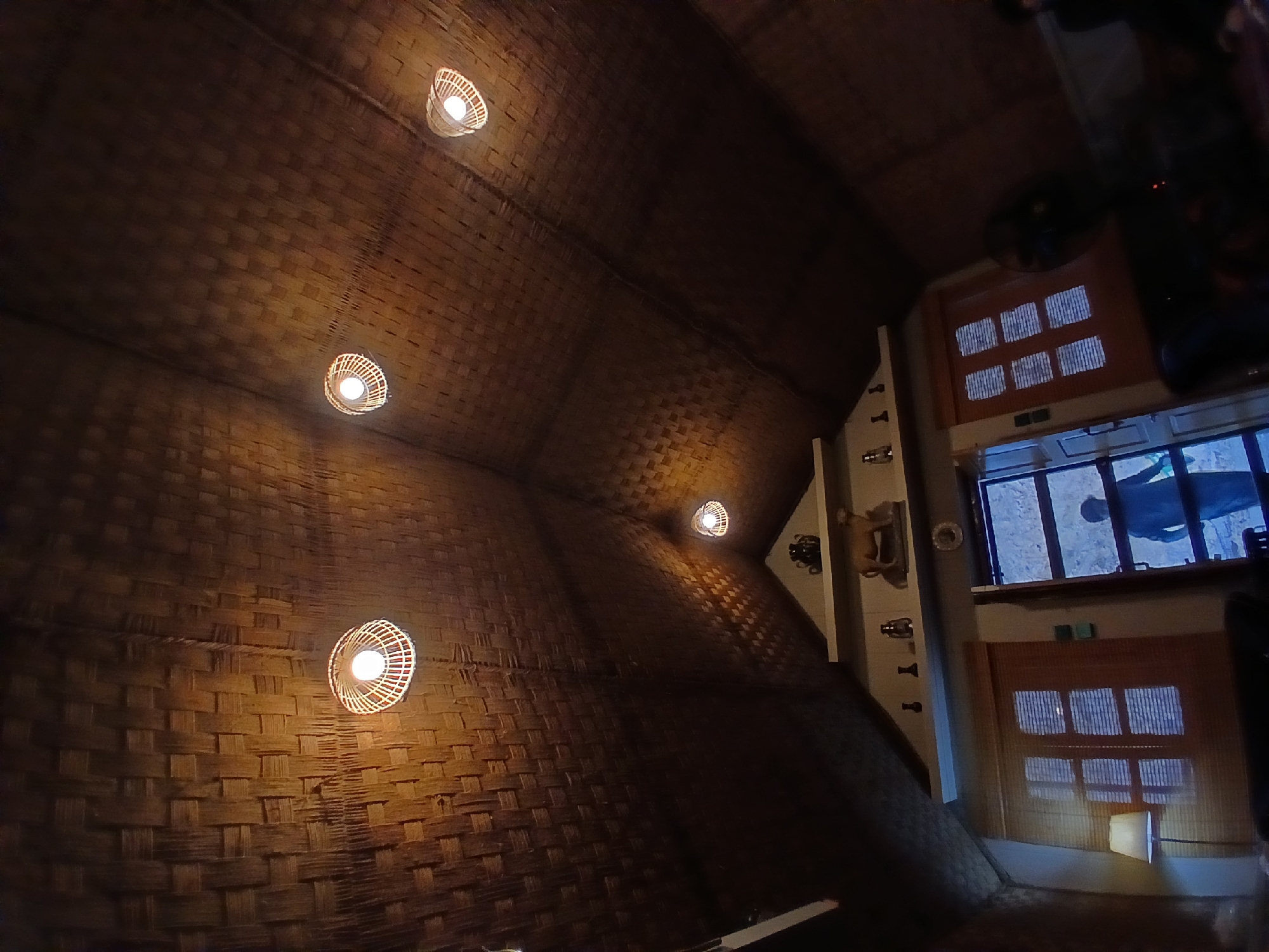The image displays the interior of a room, notable for its distinctive ceiling and walls covered in woven natural fibers, likely bamboo or rattan, which exhibit a warm brown and orange hue. The space is illuminated primarily by several round, open-weave pendant lights, each housing a bright, warm-toned bulb. These fixtures are suspended from the curved, textured ceiling, casting light onto the intricate woven patterns.
Towards the right side of the frame, there's a structural element resembling a counter or service area. Above this, a large rectangular opening or window provides a view or reflection of a person, visible as a dark silhouette against a cooler, lighter background, possibly indicating an outdoor scene or another room beyond. Below this main opening, several smaller, rectangular panels with a grid-like pattern are visible. One of these panels is illuminated from below by a small, square light source. To the left of this counter structure, on a lighter-colored wall, a few decorative objects or fixtures are mounted, including what appears to be an antique-style telephone or a similar ornamental item. A dark, circular object, possibly a fan or another light fixture, can be seen partially at the top right of the background.
The ambiance suggests a cozy, rustic, and inviting setting, heavily relying on natural materials and warm artificial lighting. The presence of bright interior lights contrasting with the cooler light in the background implies it is likely evening or night. No discernible text is visible within the image.