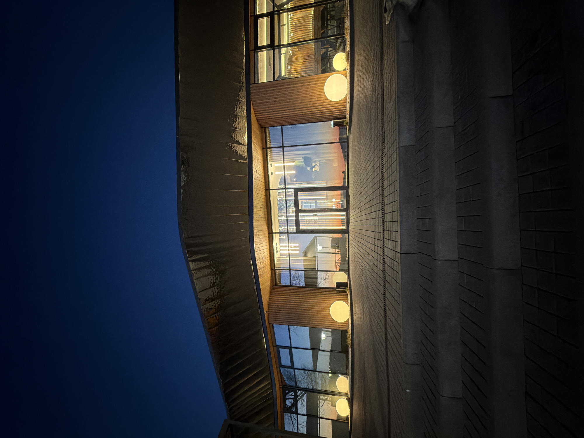 Here is a description of the image:
The image depicts a building at dusk in Votkinsk, Russia. The building features a facade with a curved, reflective metal element on the left and a series of large glass windows arranged vertically. The windows are illuminated from within, and round, glowing light fixtures are visible both inside and outside the building. The building's entryway appears to be made of glass doors and is centrally located. To the right of the building's facade, there is a stepped, dark-colored brick or concrete structure. The sky is a deep blue, indicative of twilight or evening conditions. The architecture of the building has a modern and somewhat unique design.
