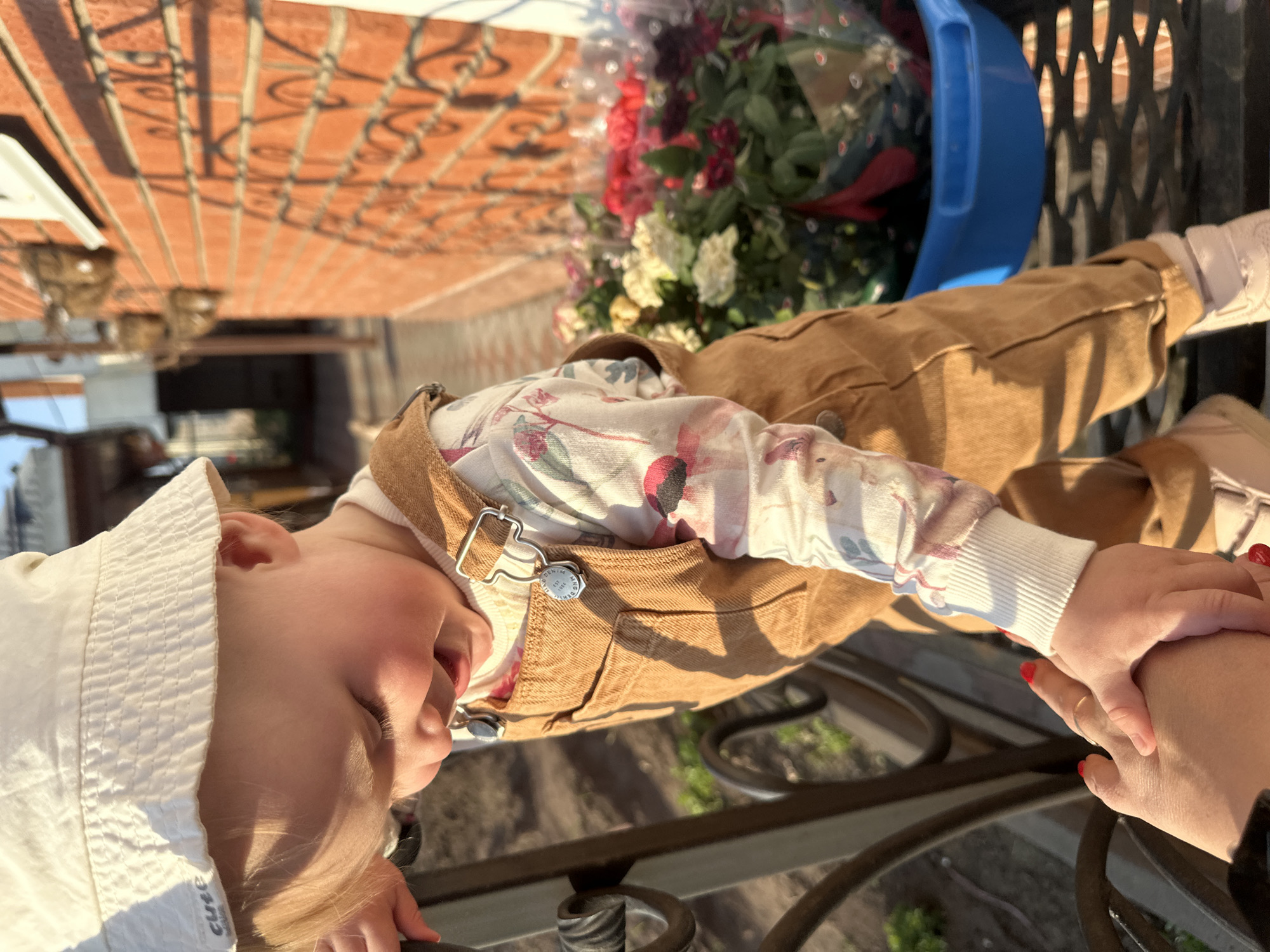 A young child lies on an outdoor metal table, holding hands with an adult. The child wears a white sun hat, a floral-patterned shirt with white ribbed cuffs, and brown overalls. The adult's hands show red nail polish and a wedding ring on the ring finger. Behind the child, a blue plastic bucket holds a bouquet of mixed flowers. The background features a brick wall with decorative metalwork. The scene is likely located in Chaikovsky, Russia, judging by the light and surroundings, though there are no explicit signs to verify this.
