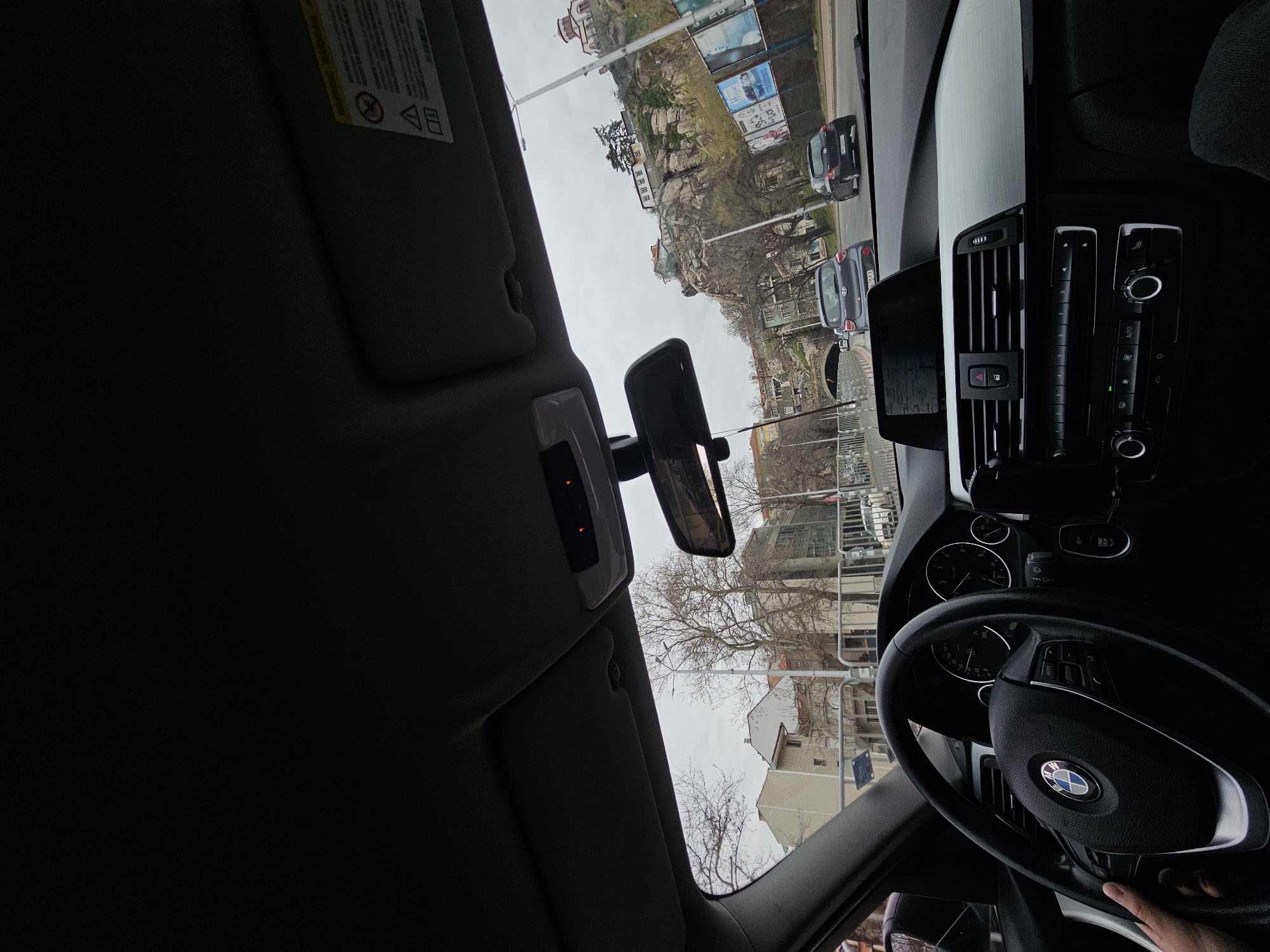 The image shows the view from inside a car, likely a BMW, in Plovdiv, Bulgaria. The dashboard and steering wheel are visible in the foreground. Through the windshield, a street scene unfolds, featuring a rock formation with buildings and billboards on top. Several cars are on the road, and buildings are visible in the background. The weather appears overcast.