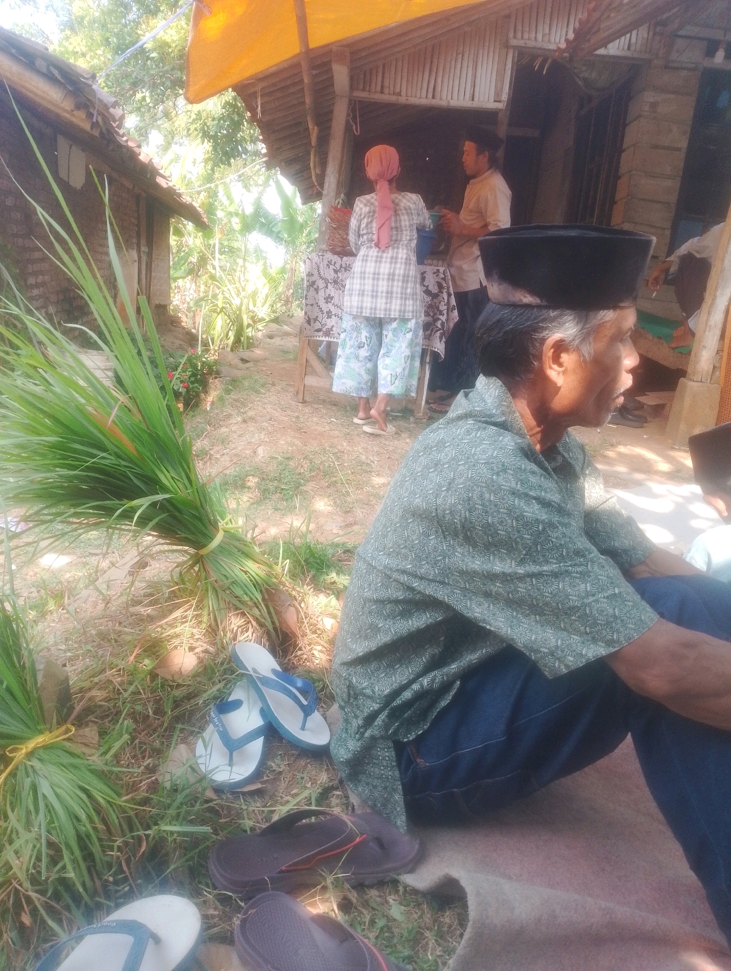 In Kalapadua, Indonesia, an outdoor scene depicts several individuals near simple structures under a yellow awning. In the foreground, an older man with grey hair wears a green patterned shirt and a black peci, seated on the ground. To his right, another person in dark clothing and a black peci is partially visible. Behind them, a woman in a checkered shirt and patterned pants, with a pink headscarf, stands near a table draped with a floral cloth. Next to her, a man in a light-colored shirt converses with her. Bunches of long green grass are tied with yellow twine and placed on the ground to the left. Several pairs of flip-flops, including white and blue ones and dark brown ones, are scattered on the grass and dirt. The environment appears to be rural, with visible vegetation and the rustic construction of the buildings suggesting a humble dwelling or gathering place.