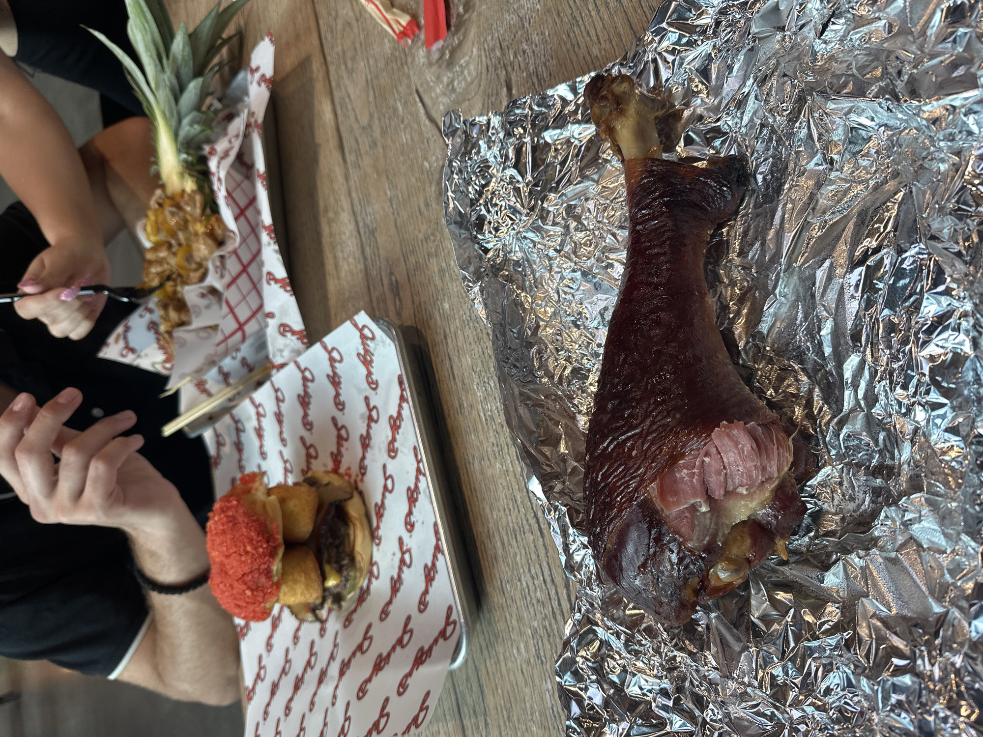 The image captures a meal on a wooden table, featuring several food items. There's a pineapple with grilled fruit chunks on a paper-lined tray. Next to it is a tray containing a burger-like item. A large turkey leg, partly consumed, rests on aluminum foil. Two people are partly visible, one using a fork to pick at the grilled fruit and another with his arm resting on the table.
The trays' liners have the logo for "Burger Culture," a restaurant in Tampa, Florida, United States. The environment suggests an indoor dining setting.