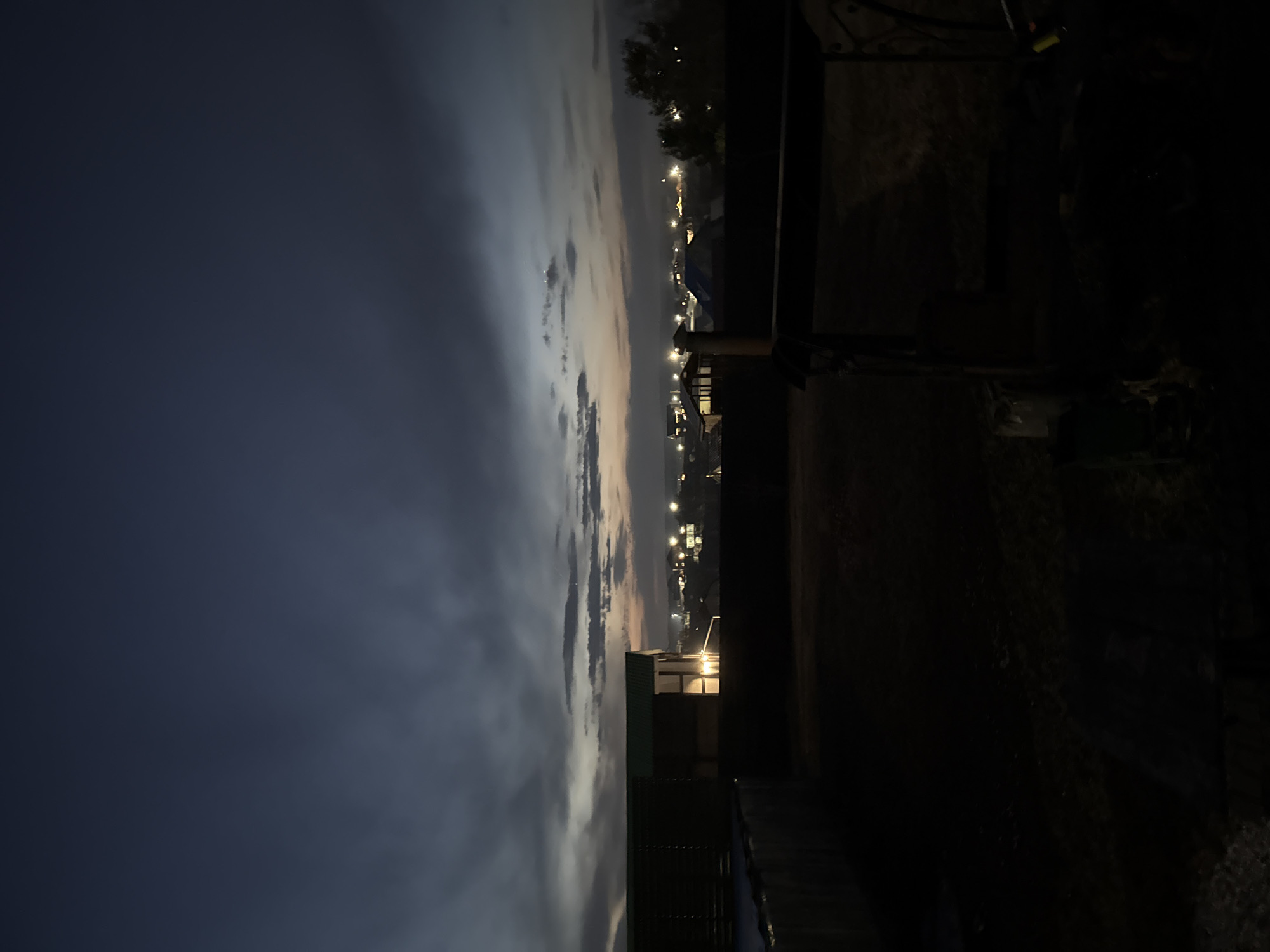 The image shows a nighttime view from what appears to be an elevated position, possibly a rooftop, in or near Бугачёво, Russia. The sky is dark blue transitioning to a lighter, cloud-filled band near the horizon where the city lights are visible. These lights suggest residential or commercial buildings. In the foreground, parts of a dark, shadowed structure obscure the view, possibly fencing or a building's upper level. The overall impression is a quiet, urban landscape at twilight.