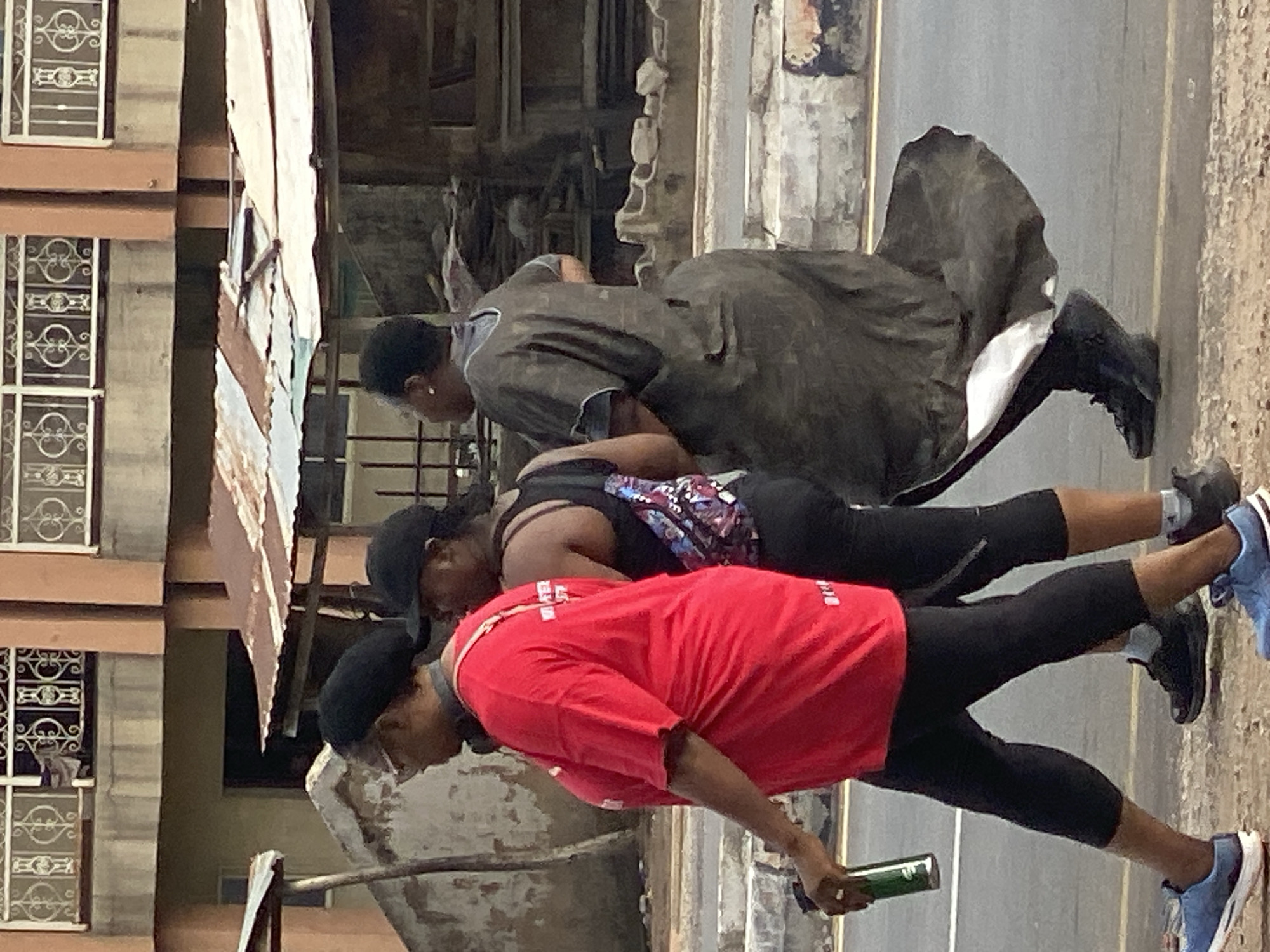 Three individuals, likely women, are captured performing a physical exercise outdoors in Adankolo, Nigeria. The image is oriented vertically, showing the subjects as if lying on their sides, with the ground extending from the bottom to the top right of the frame.
The foremost person, positioned at the bottom, wears a bright red short-sleeved t-shirt, black capri-length leggings, a black baseball cap, and blue and white athletic shoes. They hold a green cylindrical object, possibly a water bottle, in their right hand. Above them, a second person wears a dark tank top with a colorful, patterned back, black capri-length leggings, a dark cap, and black athletic shoes. The third individual, at the top of the frame, is dressed in a dark, long-sleeved tunic-like garment over dark pants and black boots; their head is partially obscured.
All three are in a dynamic, forward-leaning stance with bent knees, appearing to perform squats, lunges, or a similar exercise. They are aligned in a row, suggesting a synchronized workout.
The setting appears to be an urban street or sidewalk. The ground is a light-colored, textured surface, likely concrete or asphalt. To the left of the individuals, a building with light-toned walls and windows featuring ornate white metal grilles is visible. Above and behind the subjects, corrugated metal sheets are present, possibly part of a makeshift structure or roofing. The scene is brightly illuminated by daylight, indicating the activity takes place during the day. White text is visible on the red t-shirt, but it is unreadable.