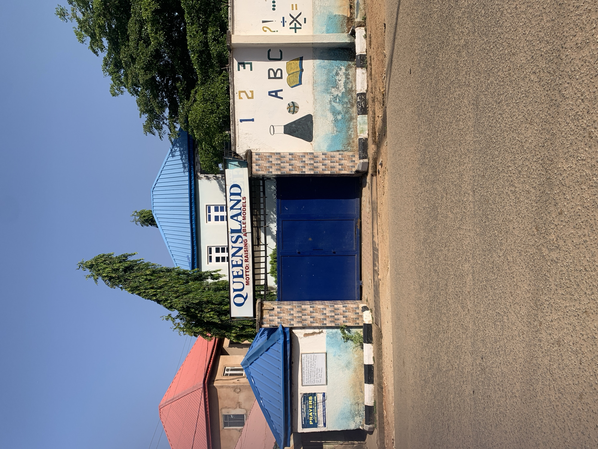 The image depicts the entrance of QueensLand School in New Stadium, Nigeria. The school's name and motto, "RAISING ABLE MODELS," are displayed prominently above the blue gate. A mural on the adjacent wall features numbers, letters, and science-related symbols. The building has a blue roof, with a red roof of an adjacent building also visible. A prayer group notice is mounted below the entrance. The street outside the school is asphalt and the sky is clear. A tall tree is visible behind the school entrance.