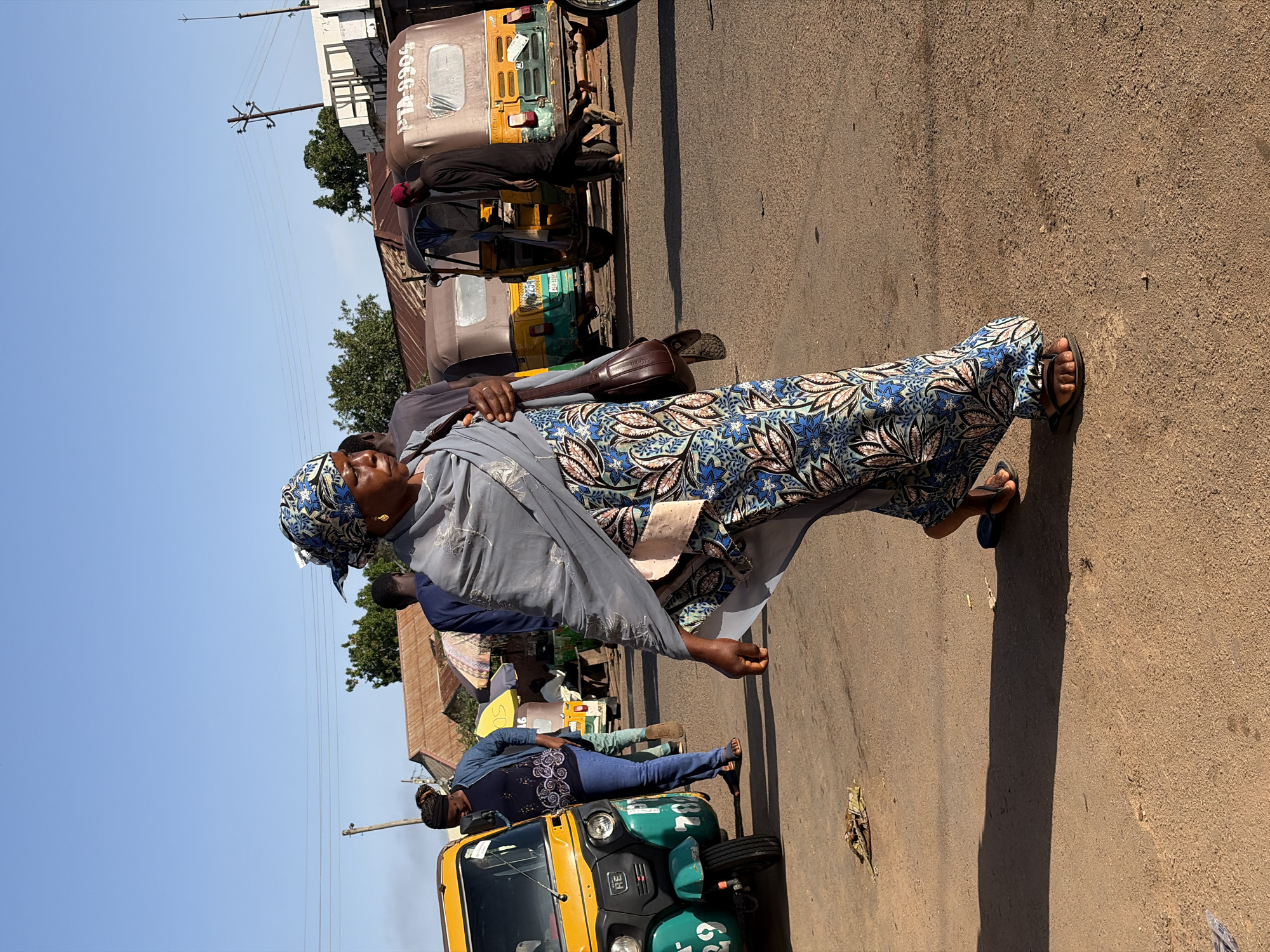 In the city of Gura Top, Nigeria, a woman walks down a dusty road. She is wearing a blue and white patterned headscarf and a grey shawl over a blue, white, and pink patterned dress. She carries a brown leather purse on her right shoulder. The road is lined with various vehicles, including a yellow and green auto-rickshaw and other auto-rickshaws in the background. Buildings with corrugated metal roofs are visible behind the vehicles. The sky is clear and blue.