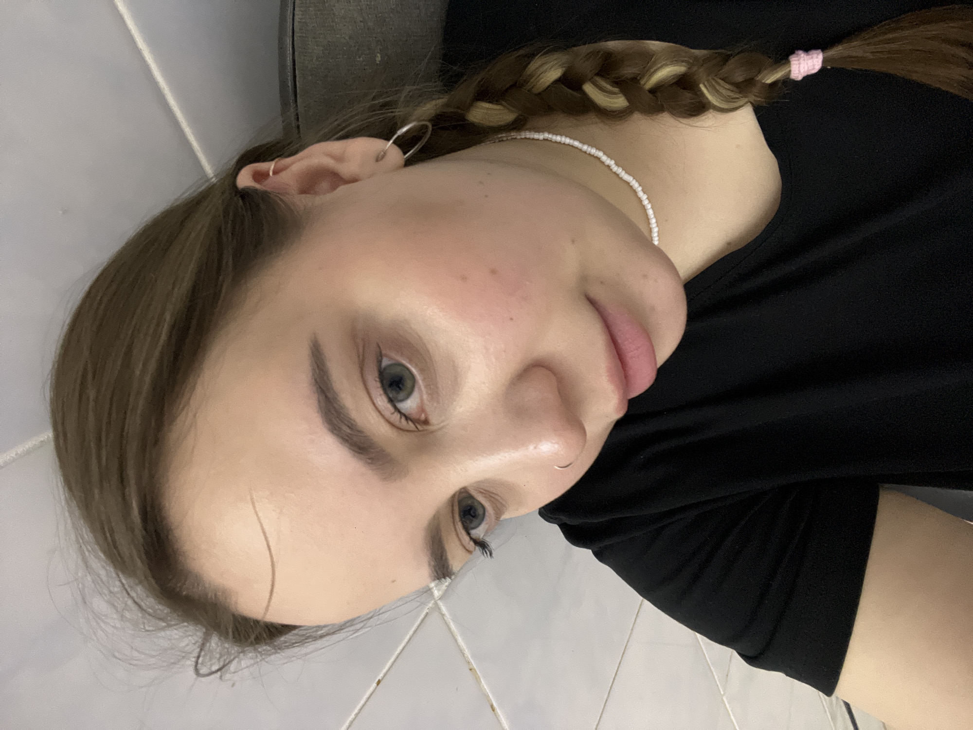 A young woman poses for a close-up selfie. She has light brown hair braided to the side with blonde highlights and secured with a pink hair tie. She is wearing a black t-shirt and a white beaded necklace. She has a nose ring and earrings. The background appears to be a tiled wall and part of a dark-colored object. There is no indication of Novopokrovskaya, Russia.