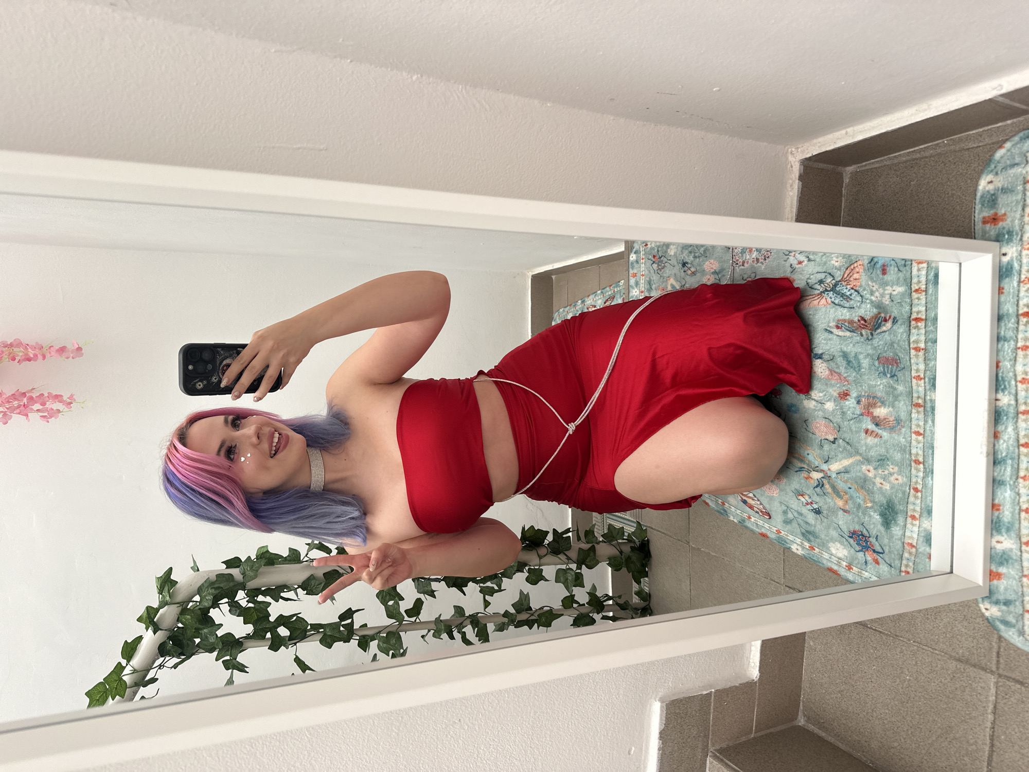 A woman with pink and blue hair is taking a selfie in a mirror. She is wearing a red two-piece outfit and a rhinestone choker. She is making a peace sign with her left hand. The mirror is leaning against a wall and has a garland of fake ivy wrapped around it. There are also some pink artificial flowers visible in the mirror. The floor is tiled and a blue rug is partially visible in the mirror. The location is not identifiable as City of Zagreb, Croatia, based on the available visual information.