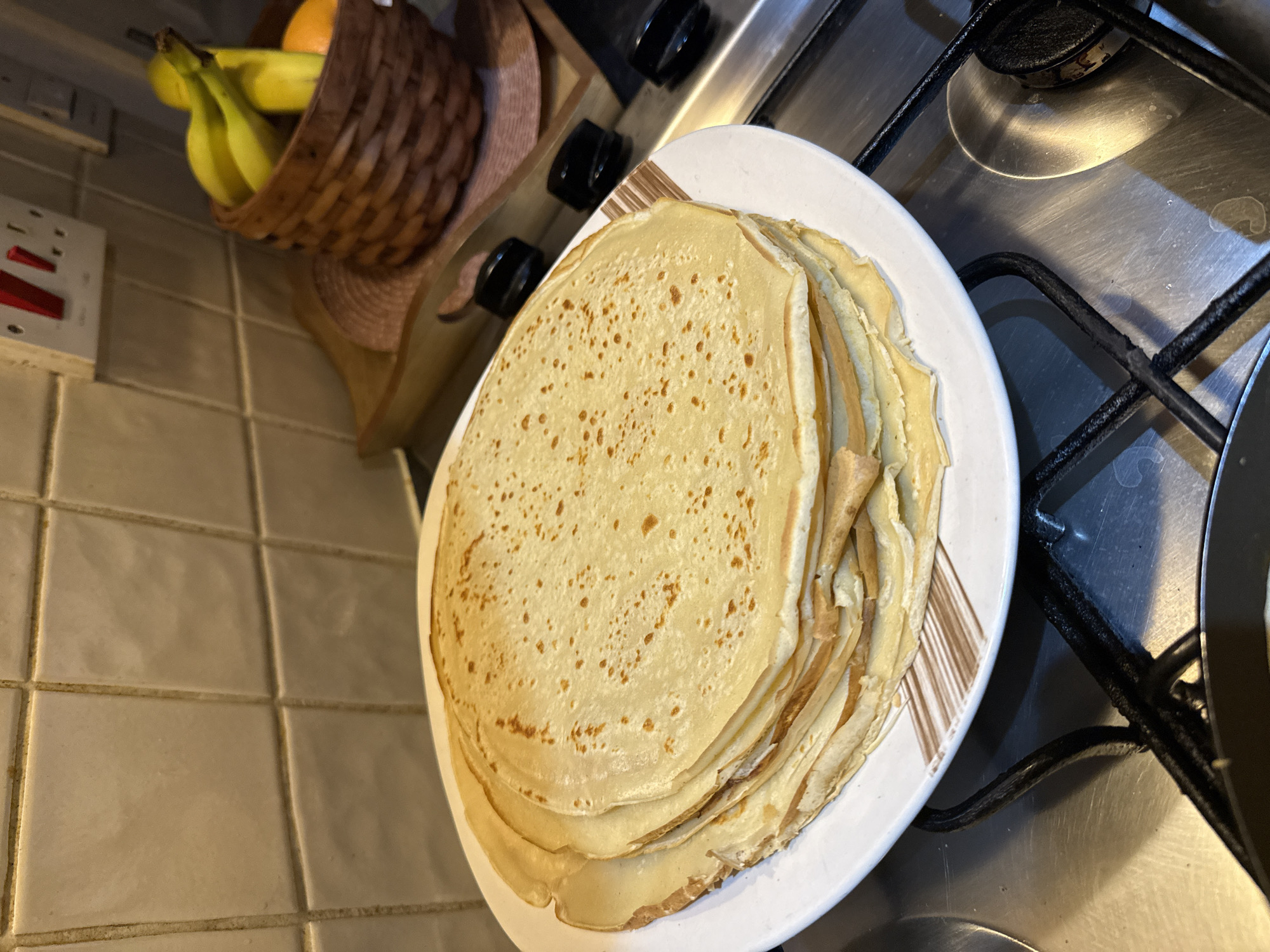 A stack of crepes sits on a white plate with a brown patterned rim, placed on a stainless steel stovetop with black grates. The stovetop also features black knobs and what appears to be a frying pan in the bottom right corner. In the background to the left, a basket filled with bananas and an orange sits atop a counter covered in light-colored square tiles. The scene likely takes place in a domestic kitchen. The image's location and city are not discernible from the provided information.
