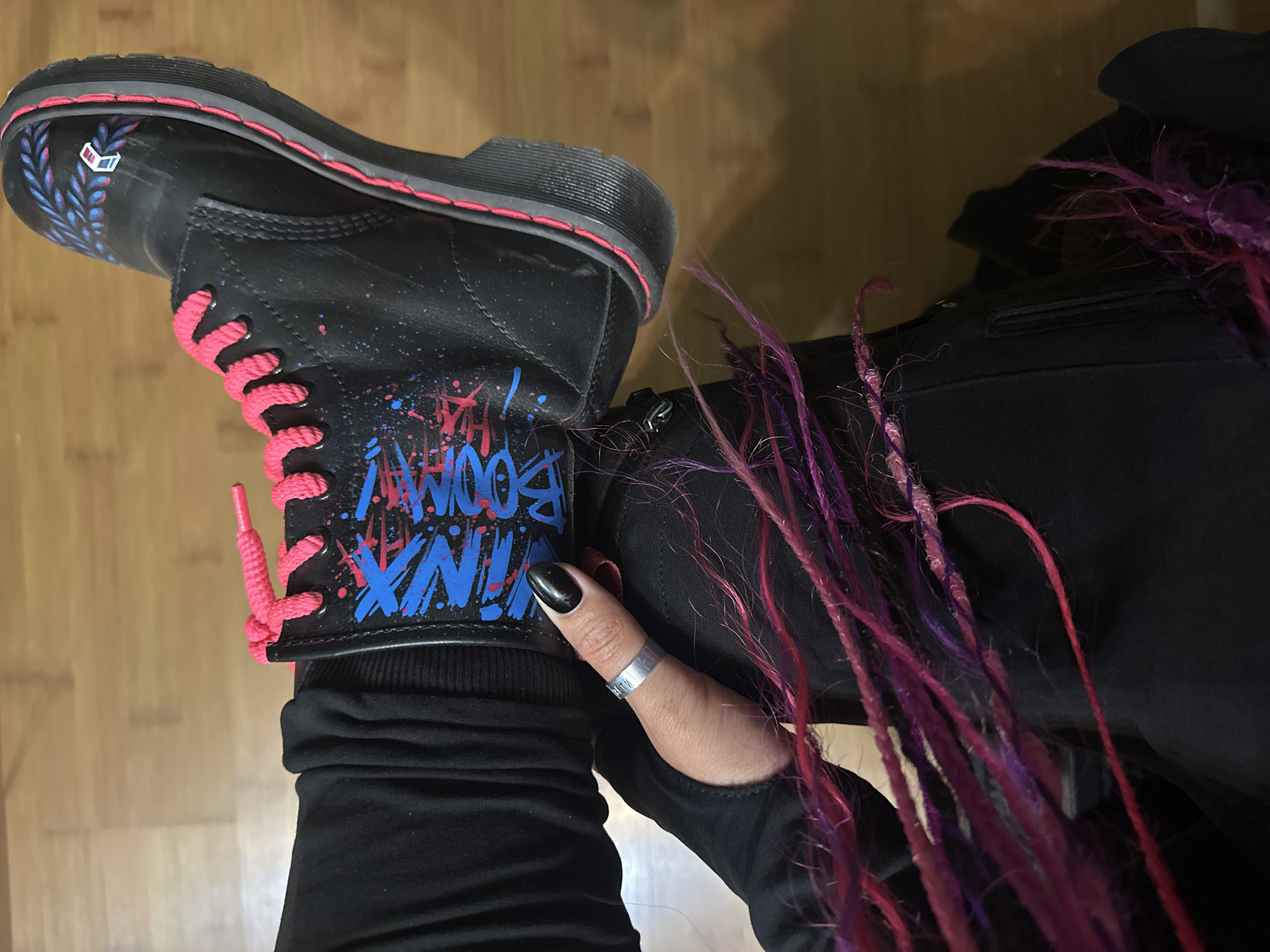 A close-up, eye-level shot depicts a person wearing a black boot with vibrant, graffiti-style designs in blue and pink. The boot is accented with bright pink laces. The person is wearing black pants and a long-sleeved black shirt. A silver ring is visible on their thumb, which has a black polished nail. The person has pink and purple hair extensions visible. The background consists of a wooden floor. The image contains no indication it was taken in Novosibirsk, Russia.