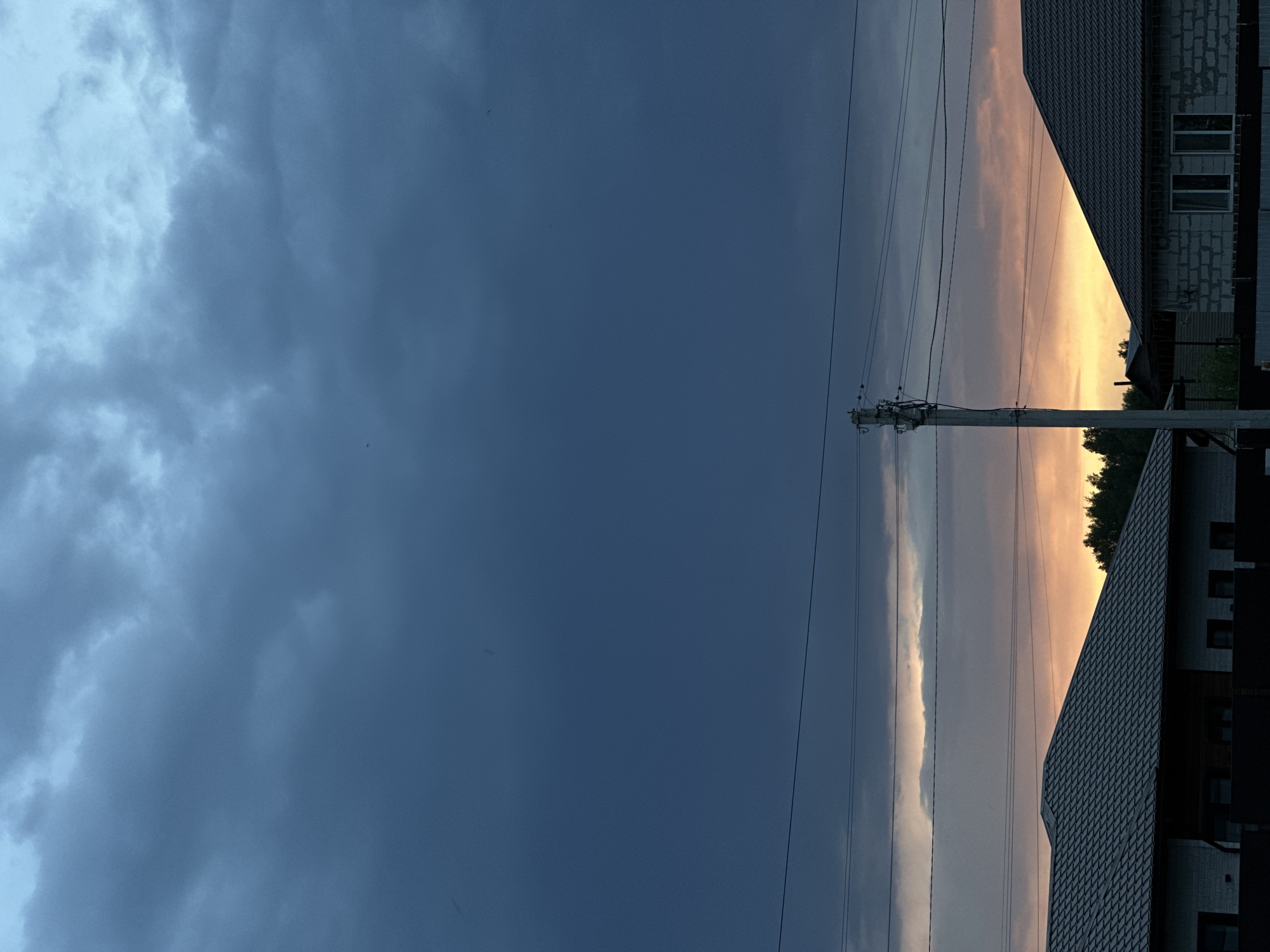 The image depicts a view of the sky and the edge of a residential area in Kharnavy, Russia. On the left side of the frame, dark storm clouds dominate the sky. To the right, there is a gap in the clouds where a sunset is visible, casting a warm orange and pink hue. Power lines run vertically through the center of the image, supported by a utility pole. The edges of houses with dark tiled roofs are visible on the right side of the frame, partially obscuring the sunset. The sky is the primary subject, showing contrasting weather conditions in different sections. The image is taken from a ground-level perspective. No people or other subjects are apparent.